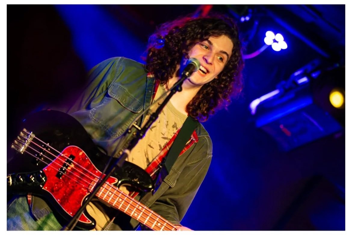A young woman with curly hair smiling while performing with an electric guitar on stage, lit by blue and purple stage lights.