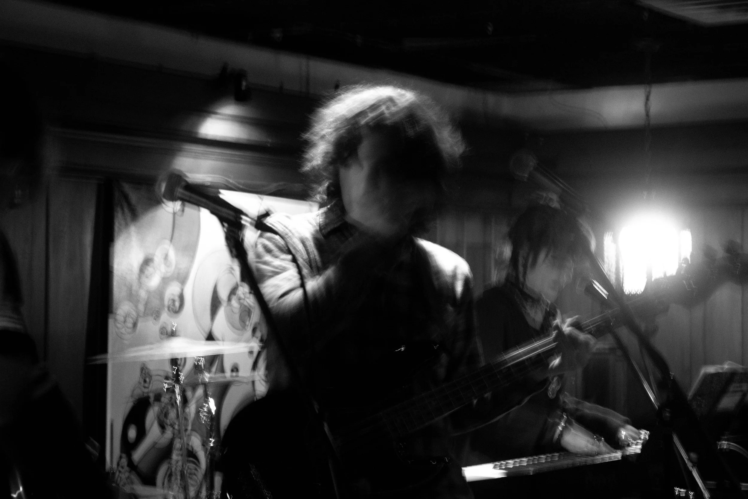 Black and white photo of two musicians performing on stage, one playing a guitar and singing into a microphone, the other playing a keyboard, with bright stage lights behind them.