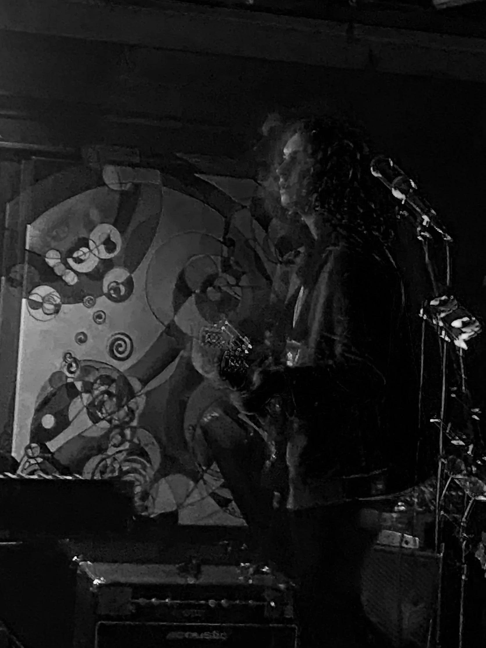 A person with curly hair playing a guitar on stage with abstract art in the background.