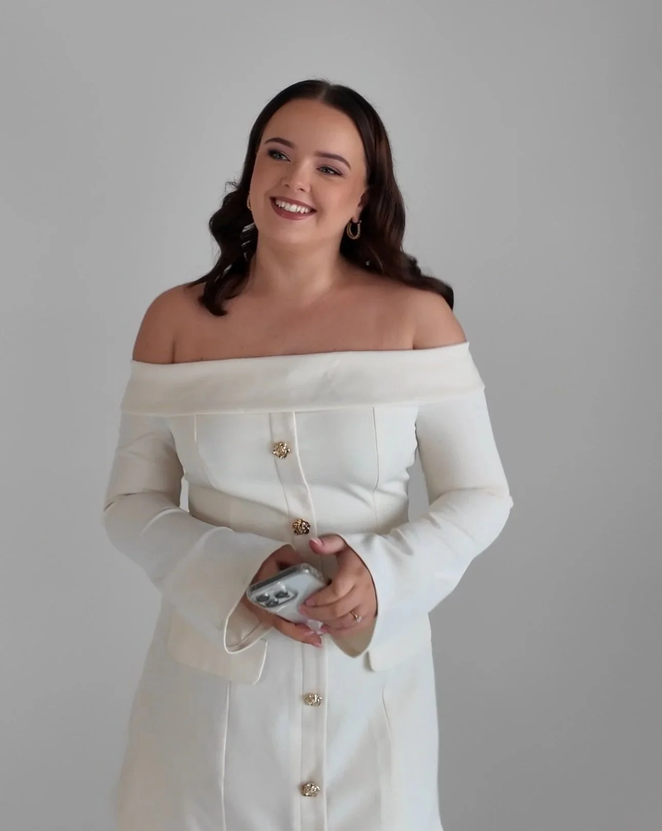 A young woman with dark hair and earrings, dressed in an off-the-shoulder white dress with gold buttons, smiling while holding a smartphone.