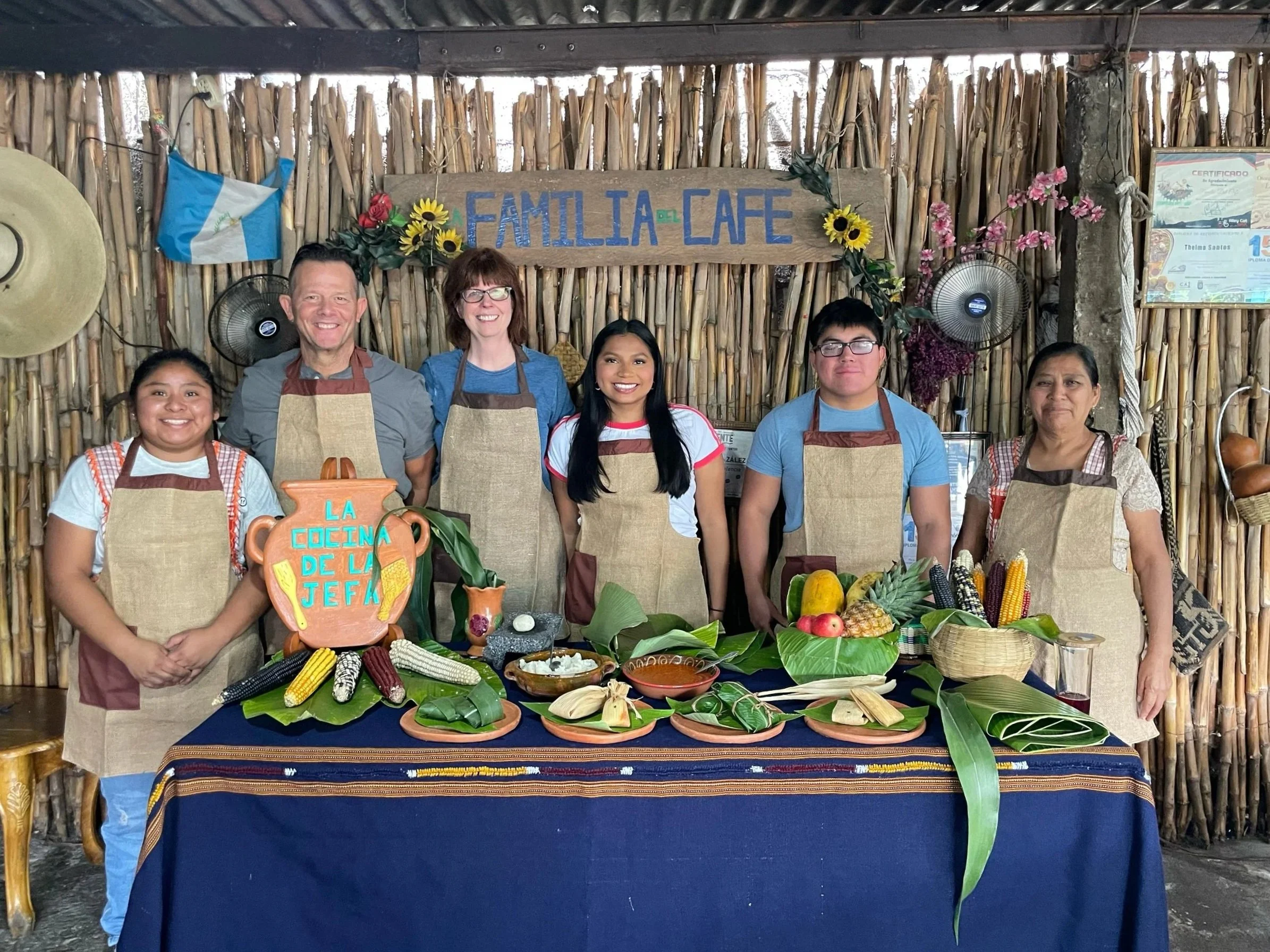 Group of seven people standing behind a table with traditional foods, in front of a rustic bamboo wall with a sign that reads 'FAMILIA CAFE'. The table is covered with a dark blue tablecloth and displays corn, fruits, and dishes. The group is wearing aprons and smiling, decorative sunflowers in the background.