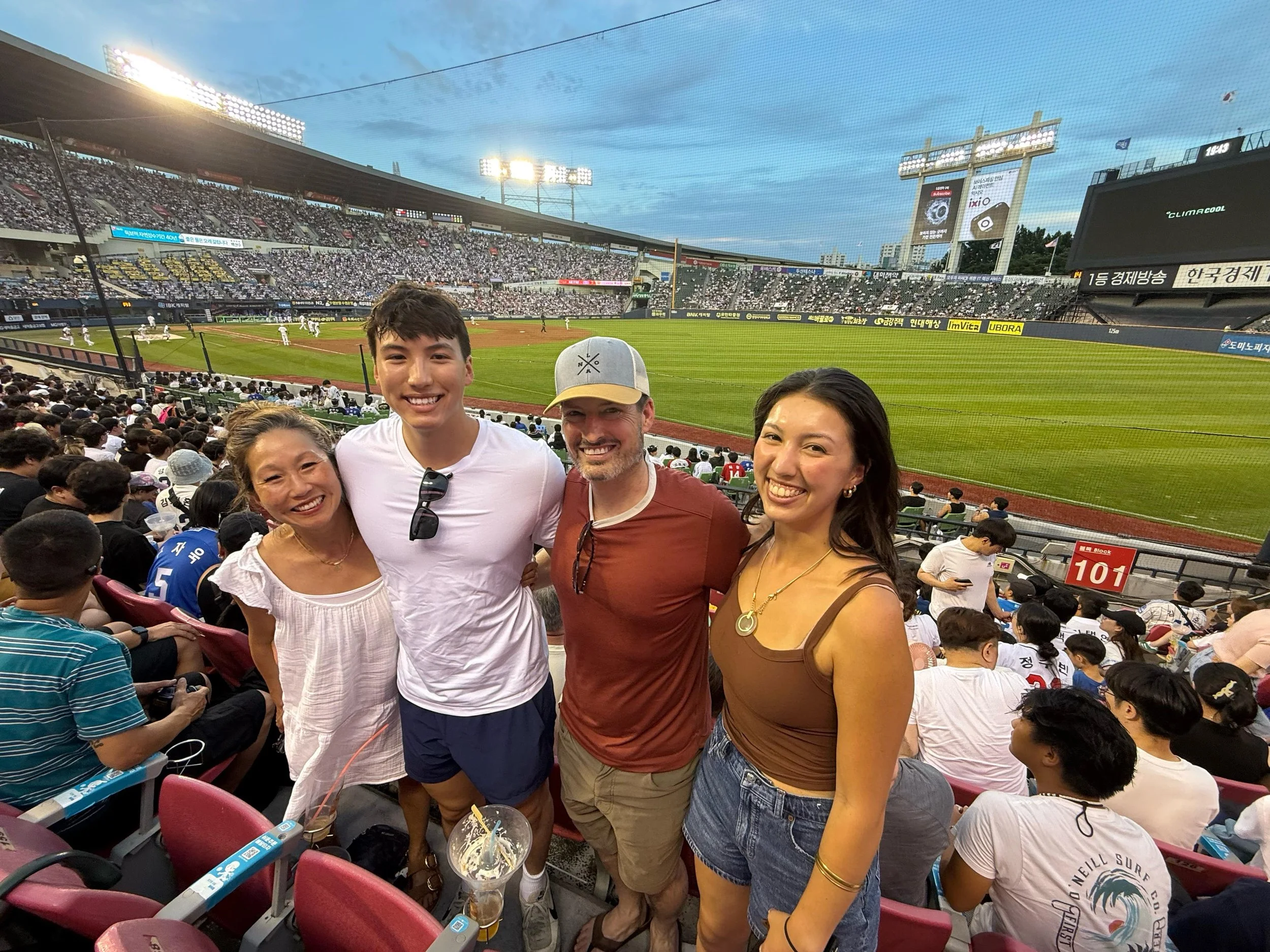 Four friends, two women and two men, smiling and posing for a photo at a baseball game in a stadium, with a baseball game in progress in the background and a crowd of spectators.