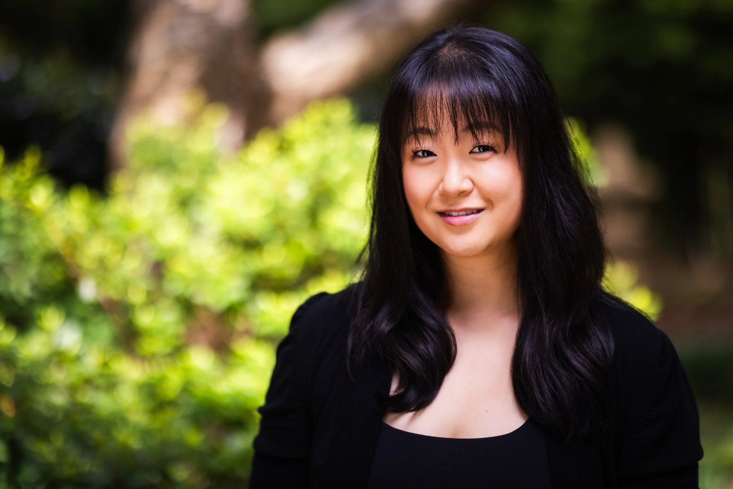 A woman with long black hair, wearing a black top and jacket, standing outdoors with green foliage in the background.
