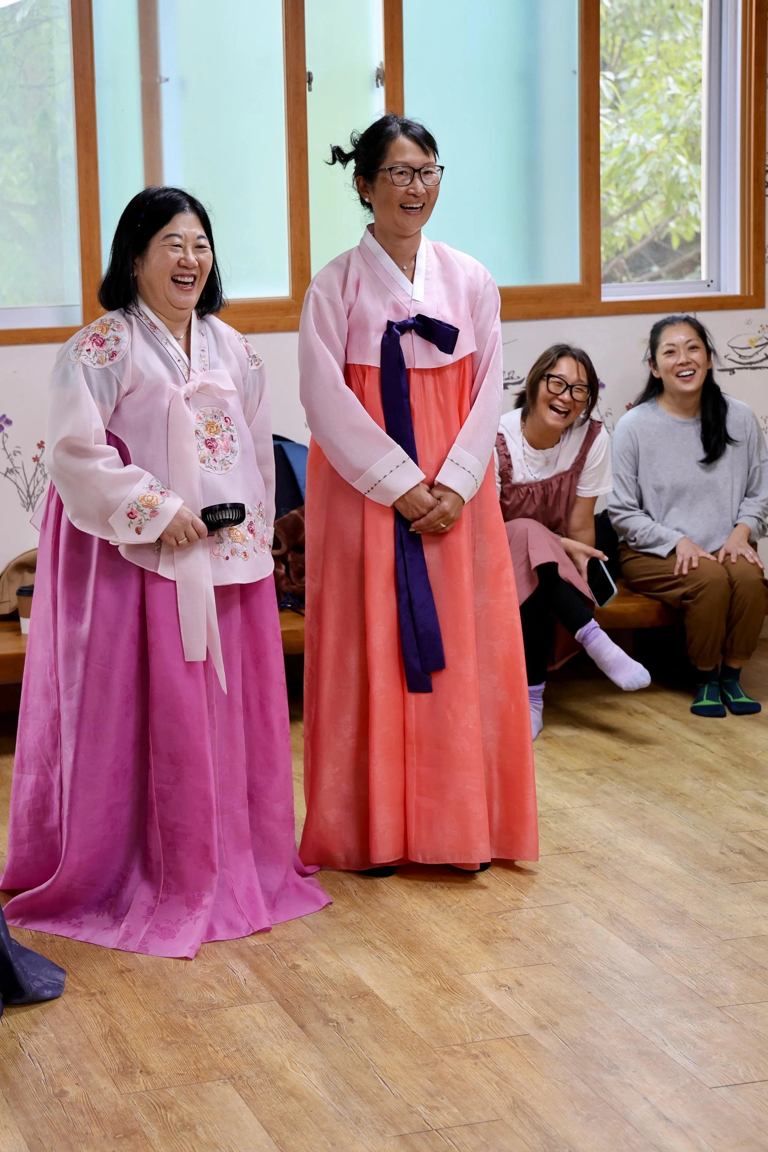 Two women in traditional Korean hanbok dresses sitting on a white sofa near a window showing cityscape view.
