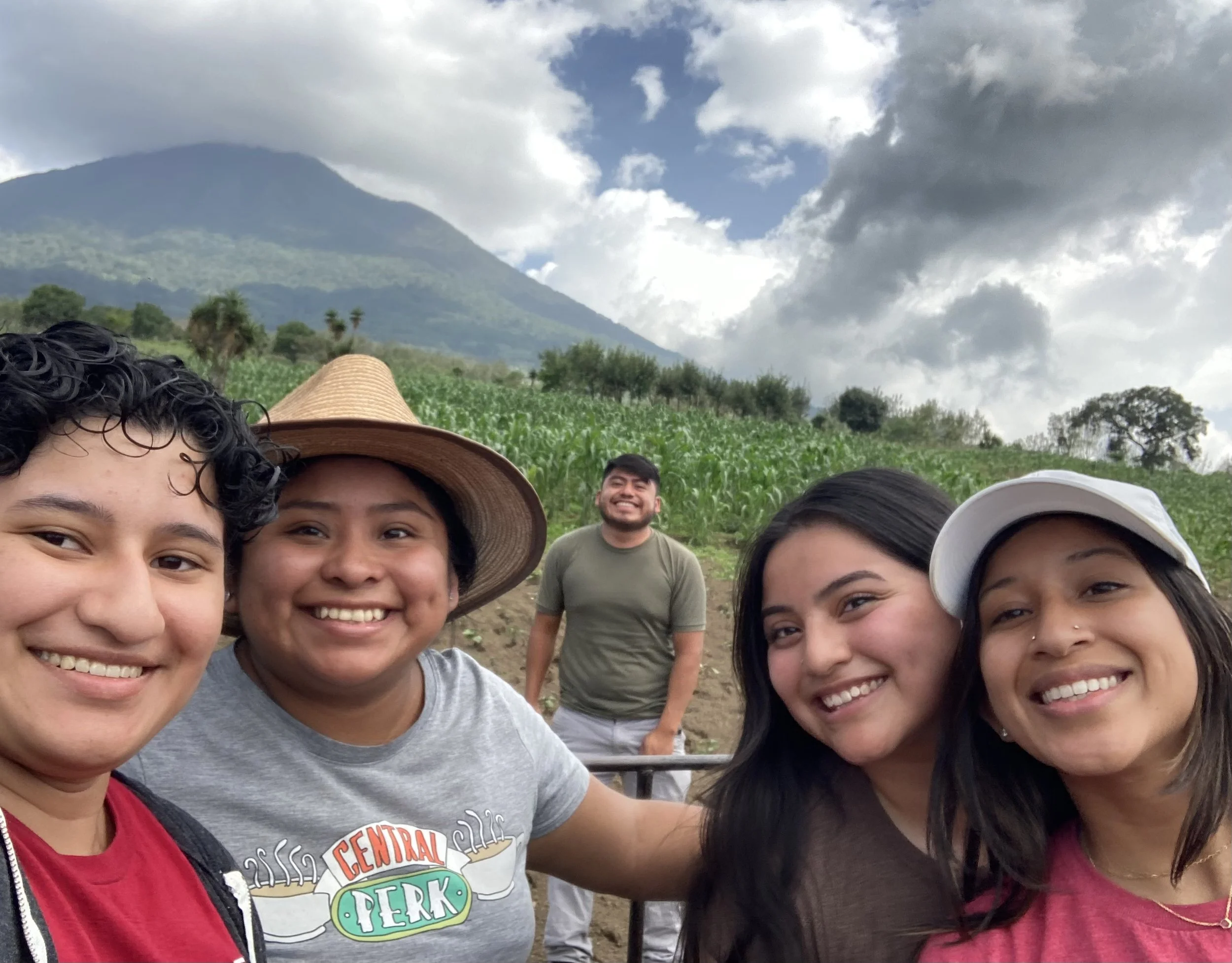 Bella and four fellow Service & Language participants smile for a selfie at La Familia del Café, a Guatemalan coffee farm, with rows of crops and a volcano in the background. Taken during Bella’s 2024 internship.