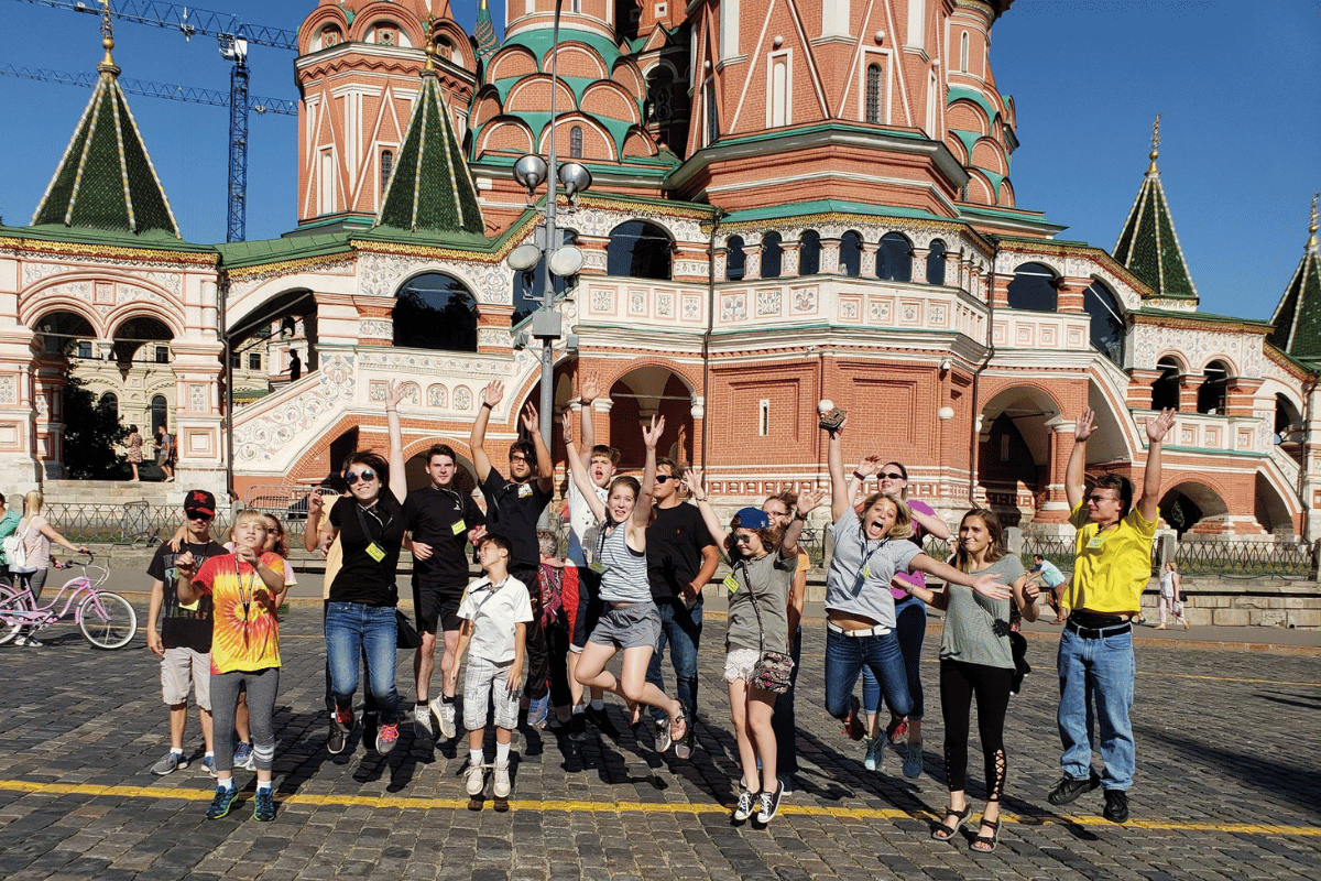 A group of children and teenagers posing playfully in front of St. Basil's Cathedral in Moscow, Russia, under a clear blue sky.