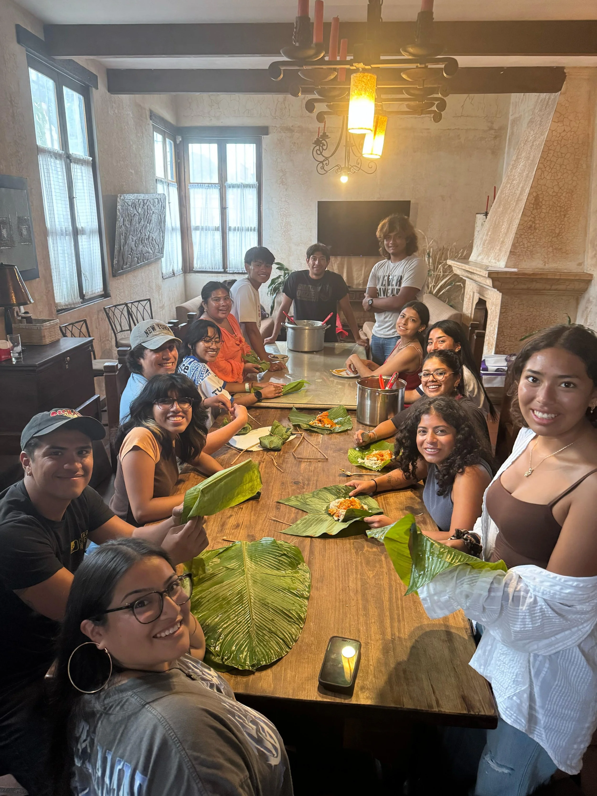 A group of adoptees gathers around a long wooden table in a warmly lit room during a cooking class in Guatemala. They’re smiling and wrapping food in large green leaves, preparing a traditional dish together.