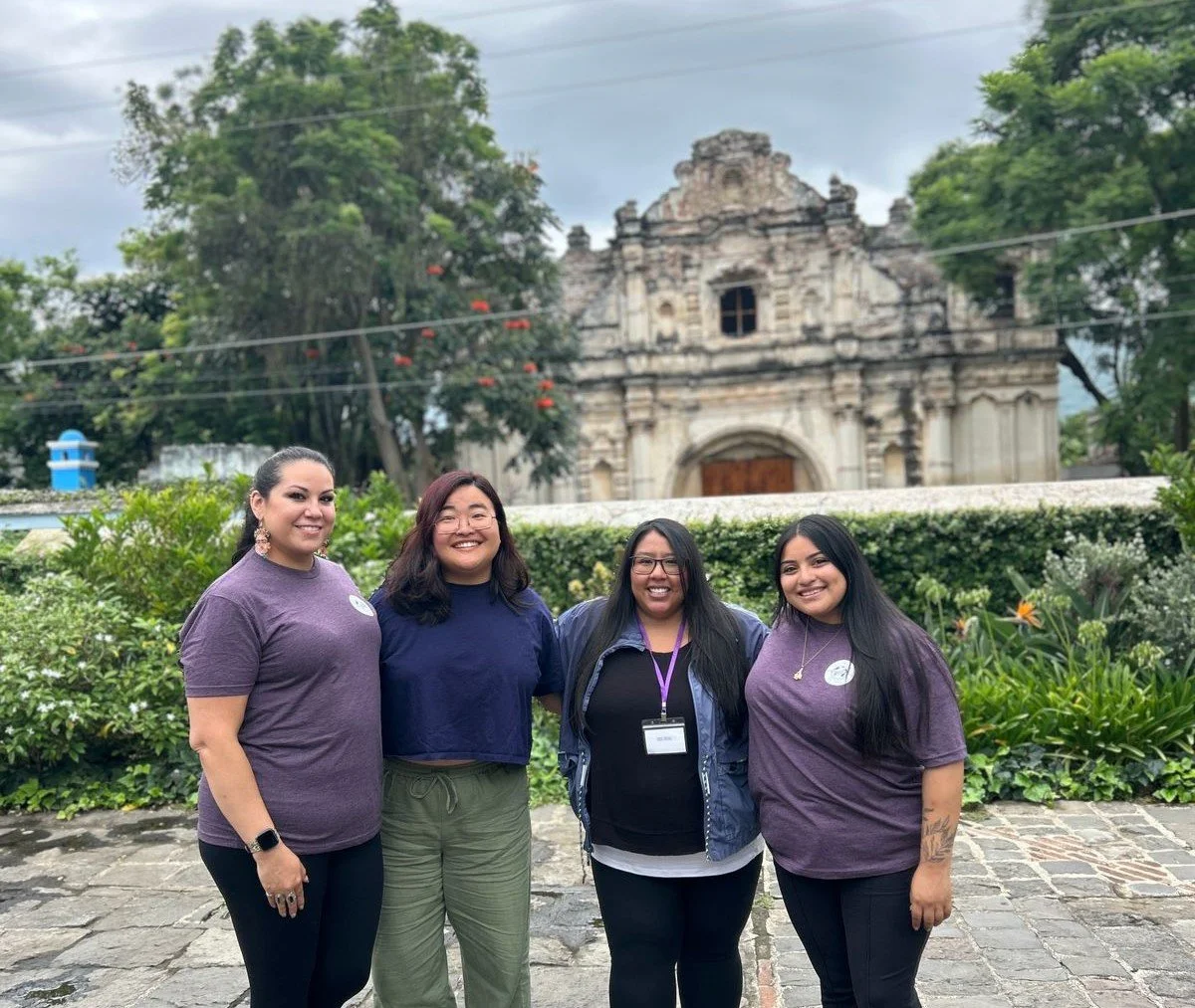 Four members of the Guatemala Ties staff smile together in front of a historic colonial church in Antigua, surrounded by lush greenery and cobblestone paths.