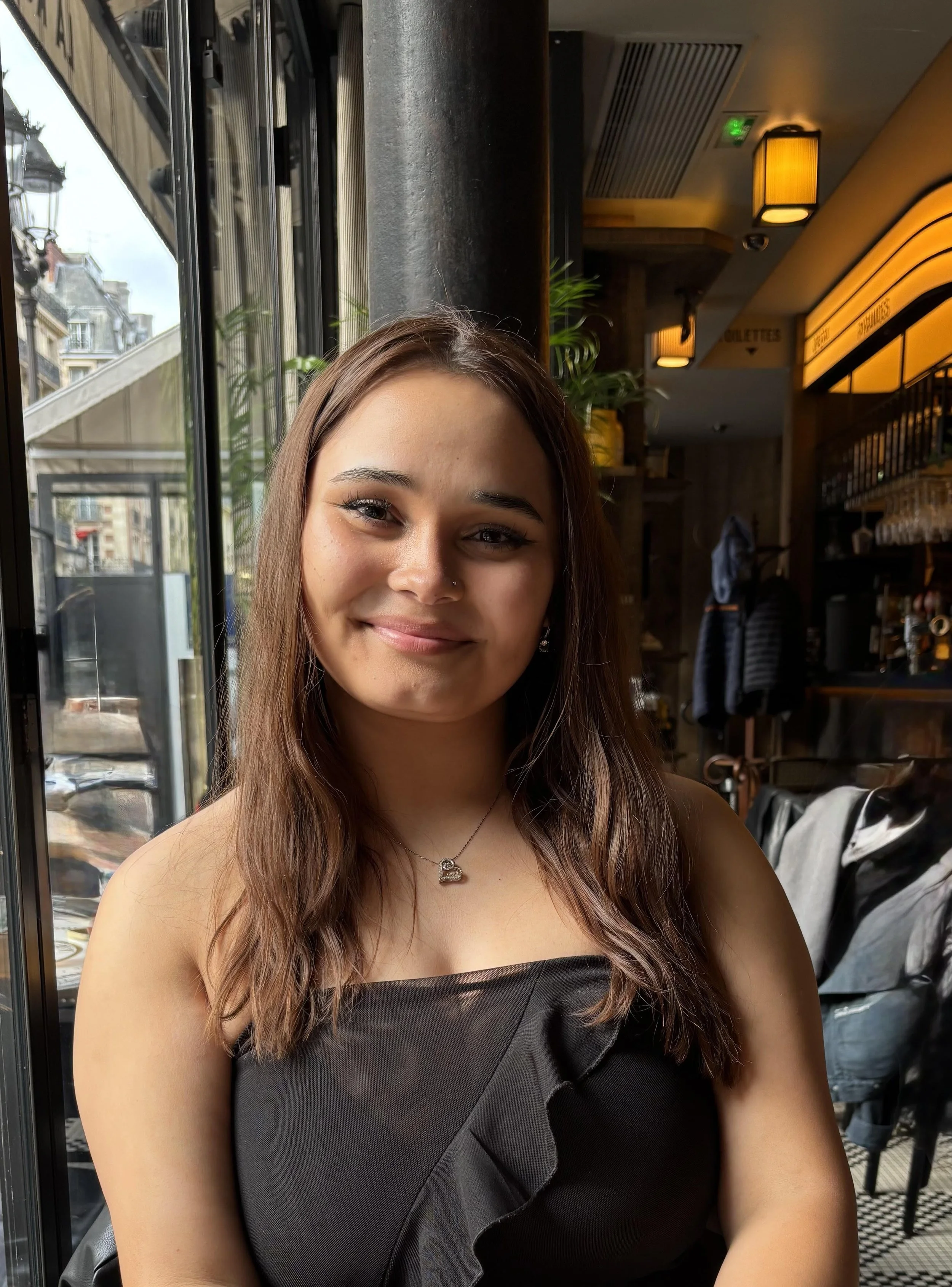 A young woman with long brown hair, smiling, sitting inside a cafe near a large window with a city street view outside. She is wearing a black strapless top with ruffled detail and a necklace.