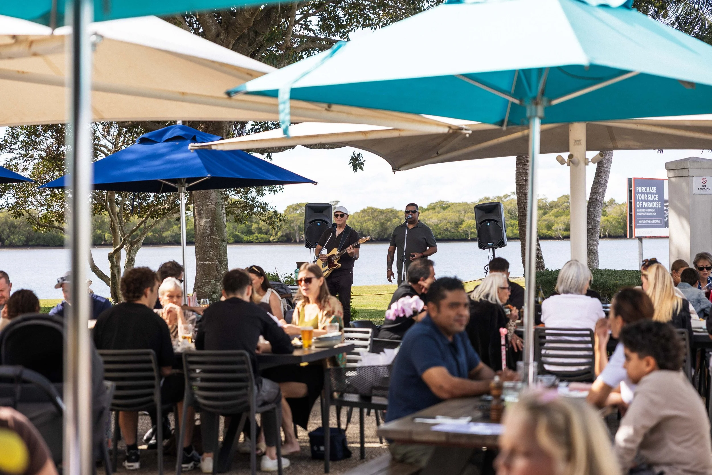 People dining outdoors under umbrellas with live music performance by two musicians, one playing guitar, near a body of water with trees in the background.
