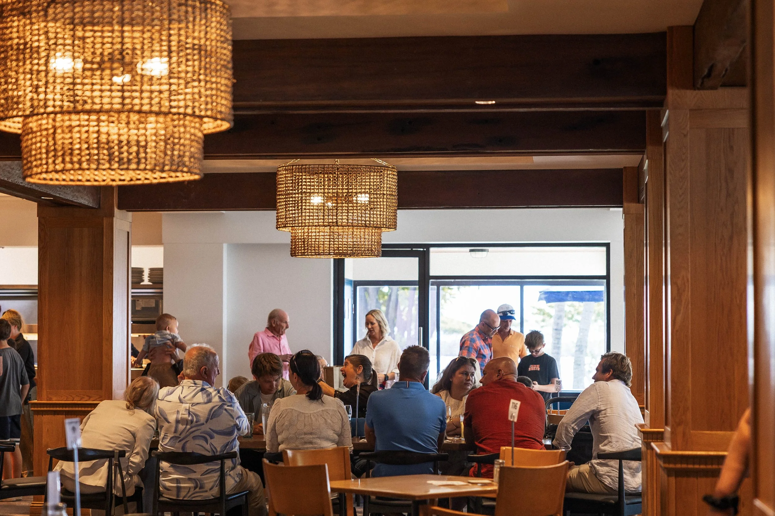 Indoor restaurant or cafe filled with people sitting at tables, talking and dining. Warm lighting from woven pendant lights hanging from a dark wooden ceiling, large window in the background letting in natural light.