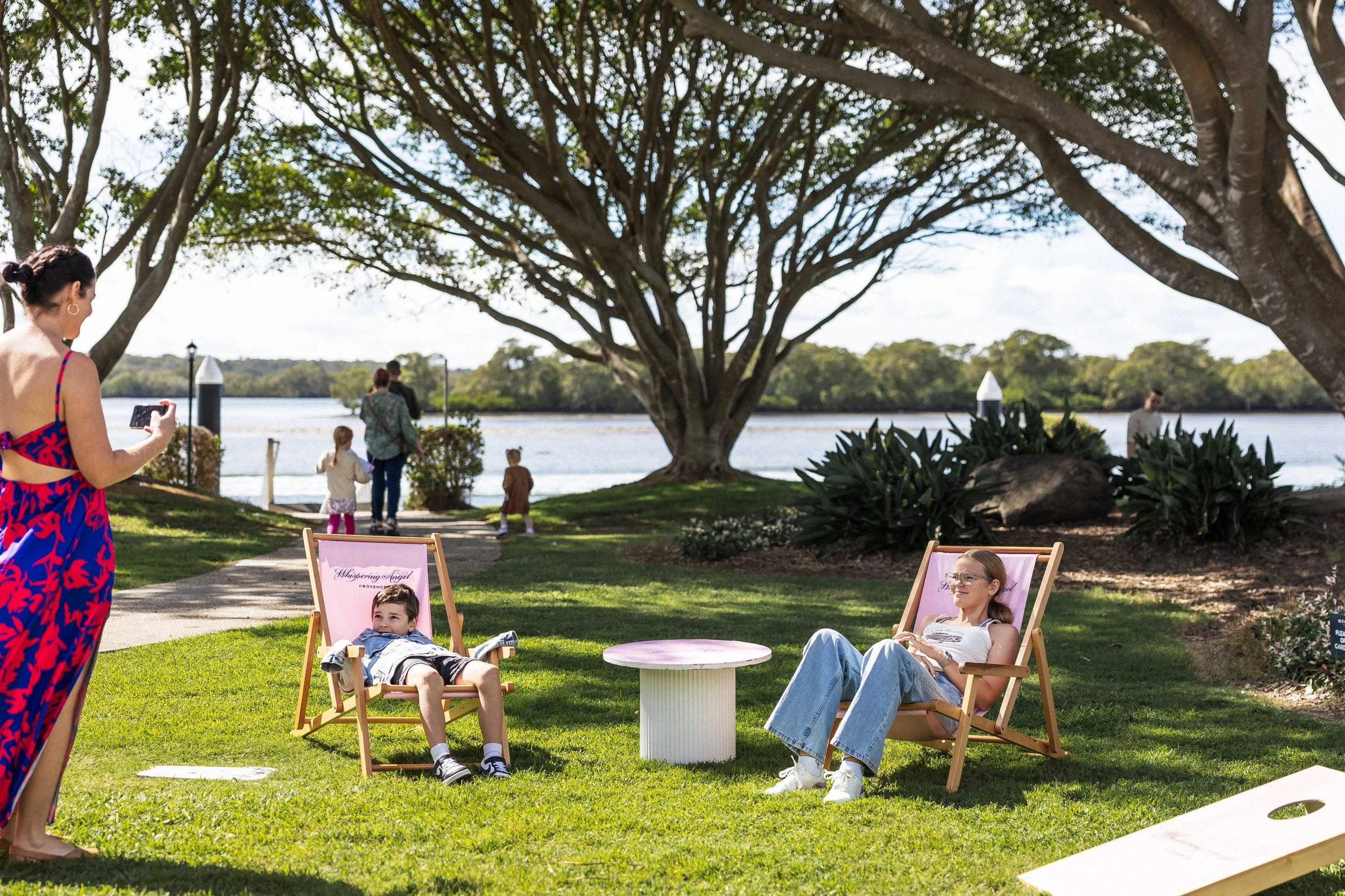 People relaxing and taking photos near a large tree by a body of water on a sunny day, with some seated in chairs and others standing and walking in the background.