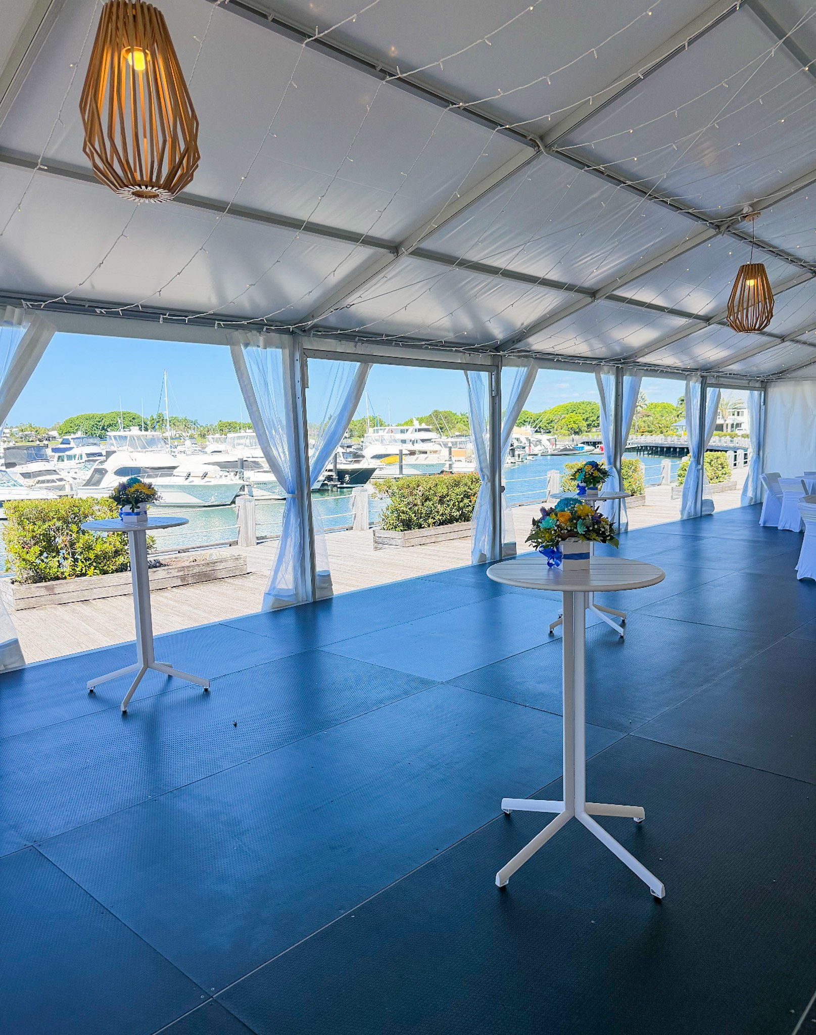 Beachside event area with blue flooring, white tables with floral centerpieces, surrounded by white curtains, overlooking boats docked at a marina on a sunny day.