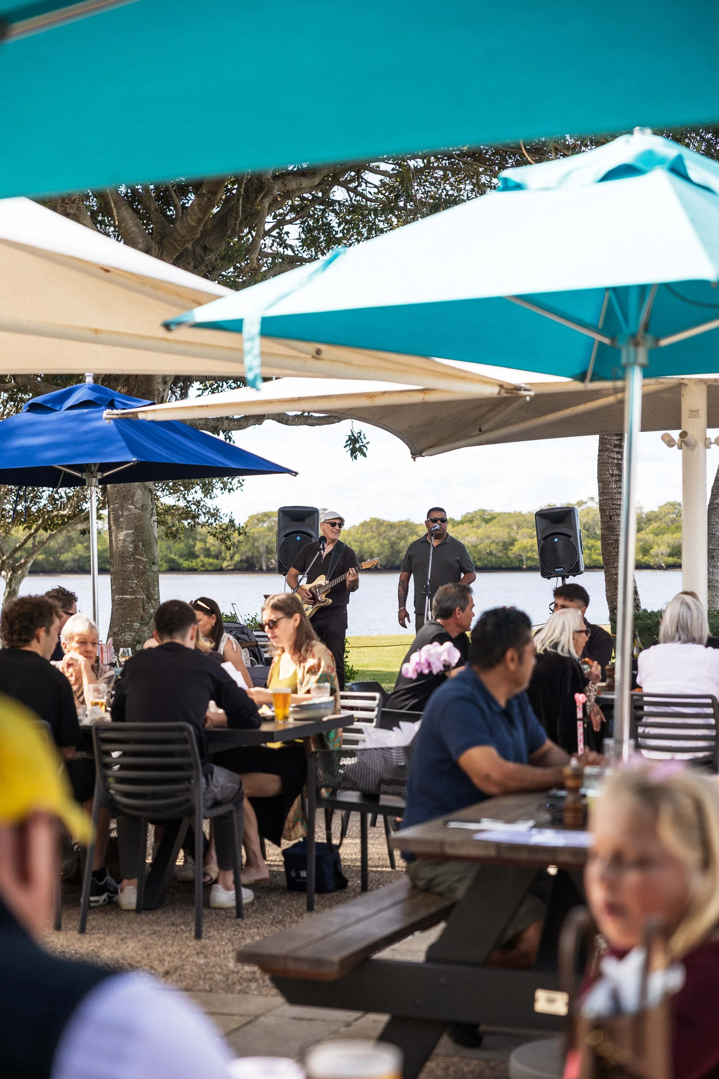 People dining outdoors under umbrellas near a body of water, with a live band performing in the background.