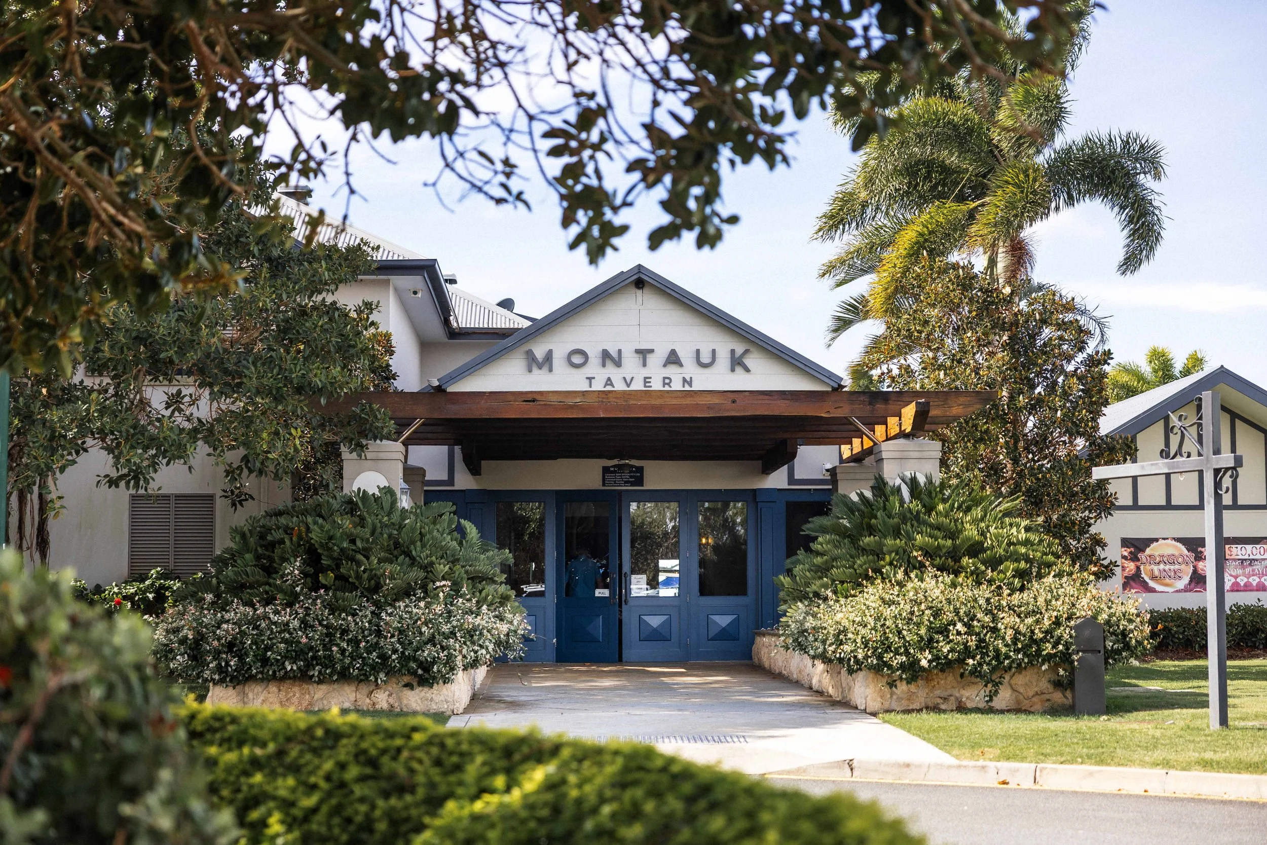 The entrance to Montauk Tavern with a blue door, surrounded by lush green bushes and trees, including palm trees, and a pathway leading up to the door.