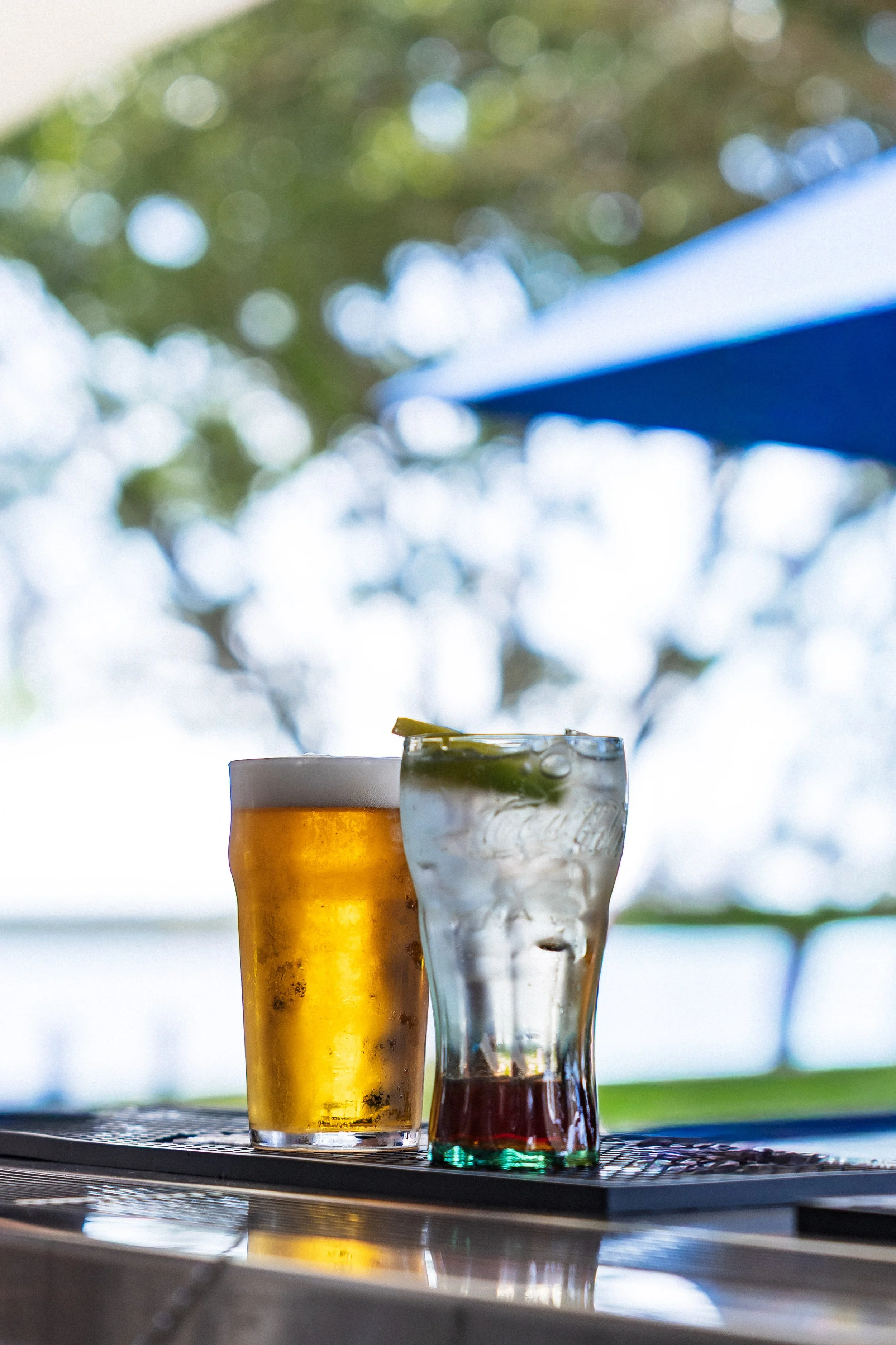 Three drinks on a bar counter with a blurred outdoor background. One appears to be beer, another a mixed drink, and the third a dark cocktail.