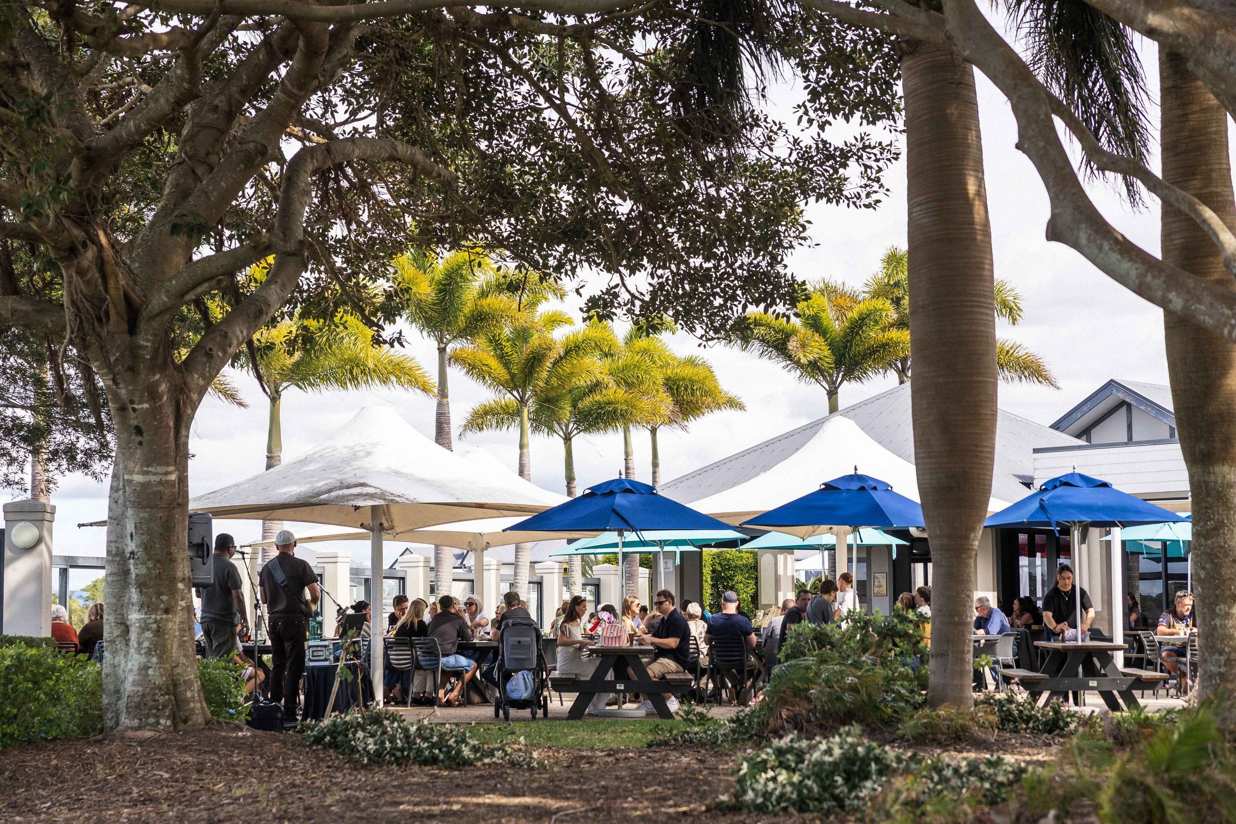 An outdoor dining area filled with people under umbrellas, surrounded by trees including palm trees and other large trees with thick branches, with a building with a gray roof in the background.