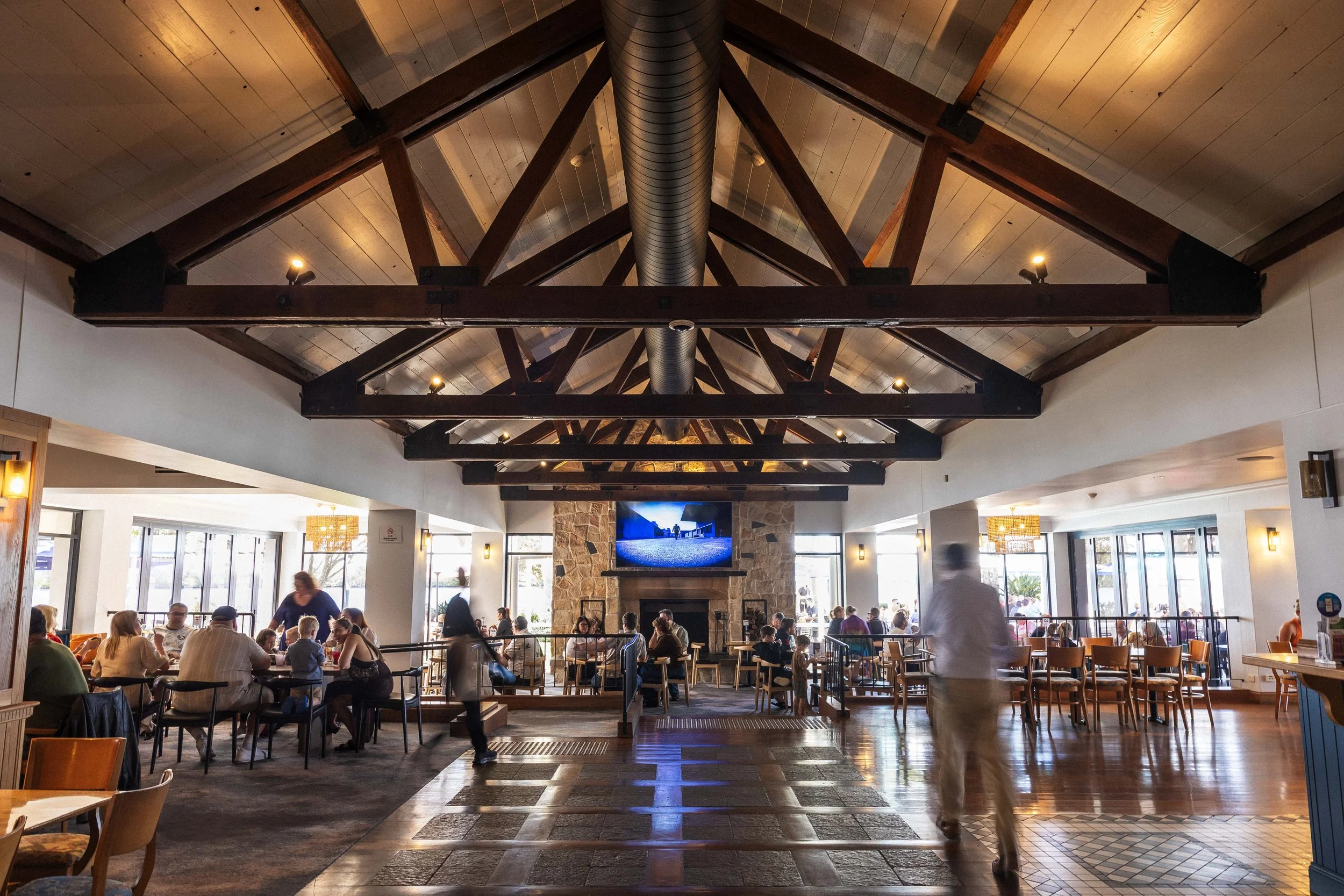 People dining and socializing inside a restaurant with a high wooden beamed ceiling, large windows, and a stone fireplace.