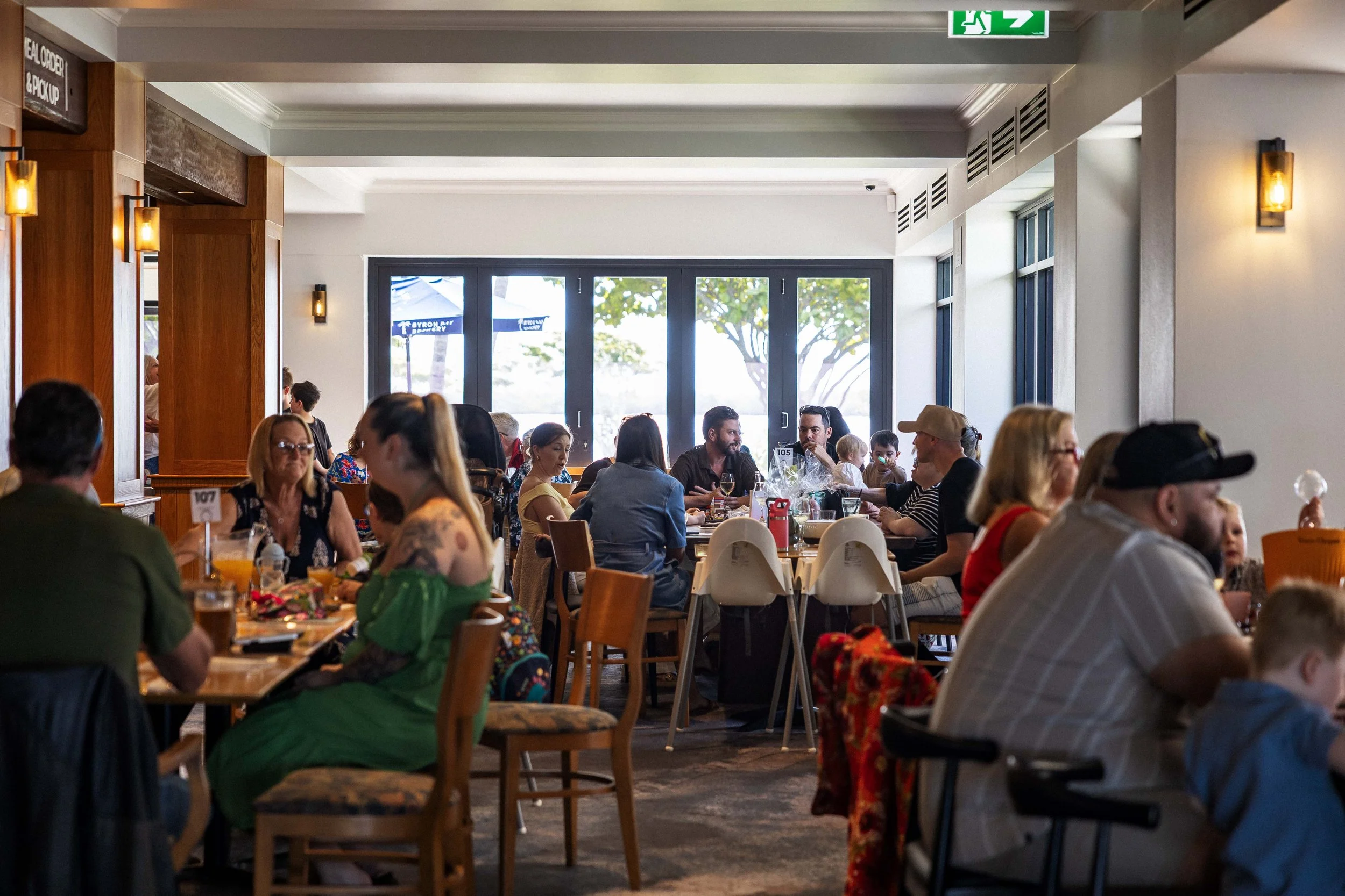 A busy restaurant interior with people sitting at tables, enjoying meals and conversation. Large glass doors open to a view of trees and outdoor umbrellas.