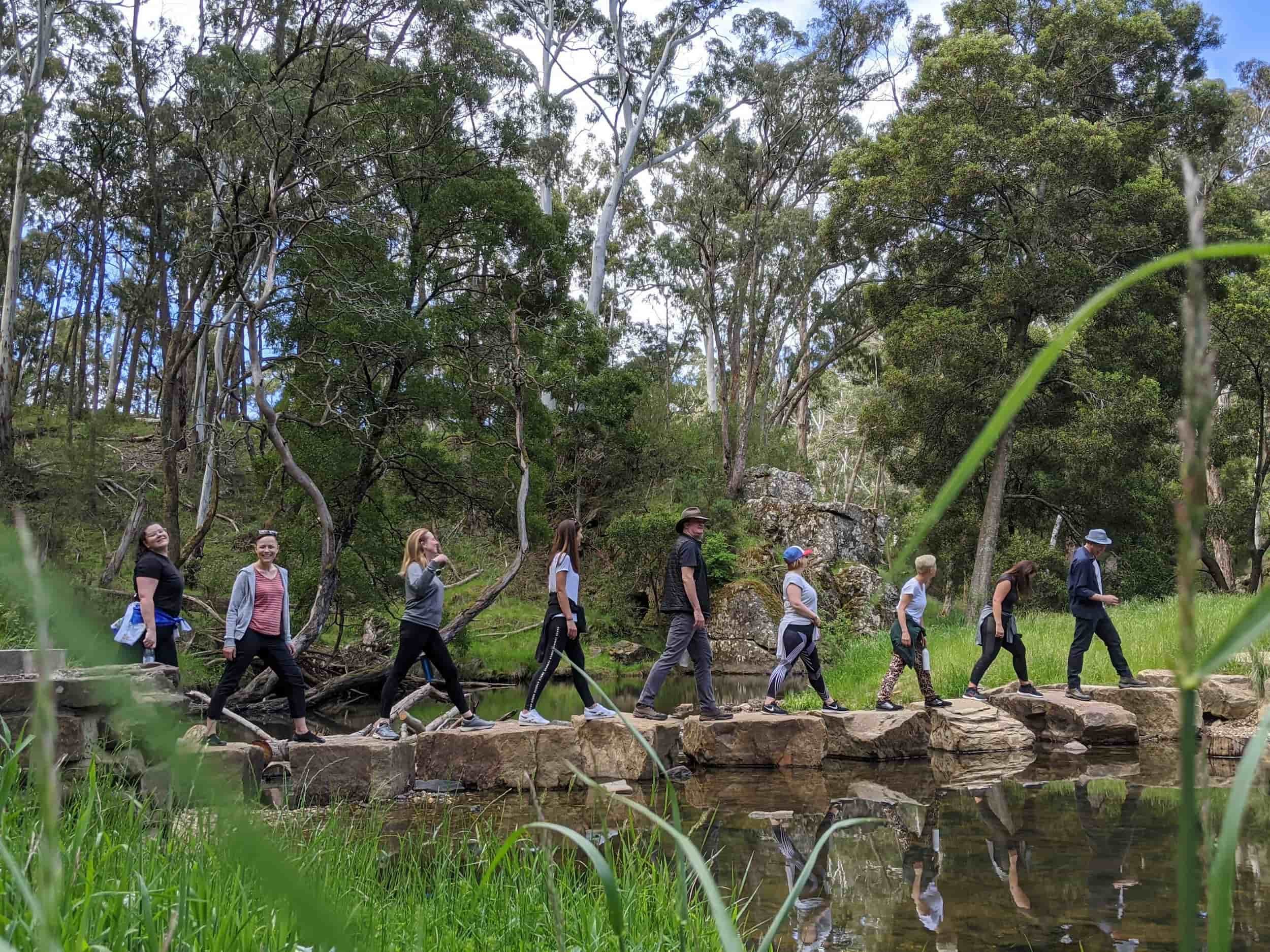 Group of nine people, including children and adults, walking on a rocky trail across a small stream in a lush green forest with tall trees and rocks.