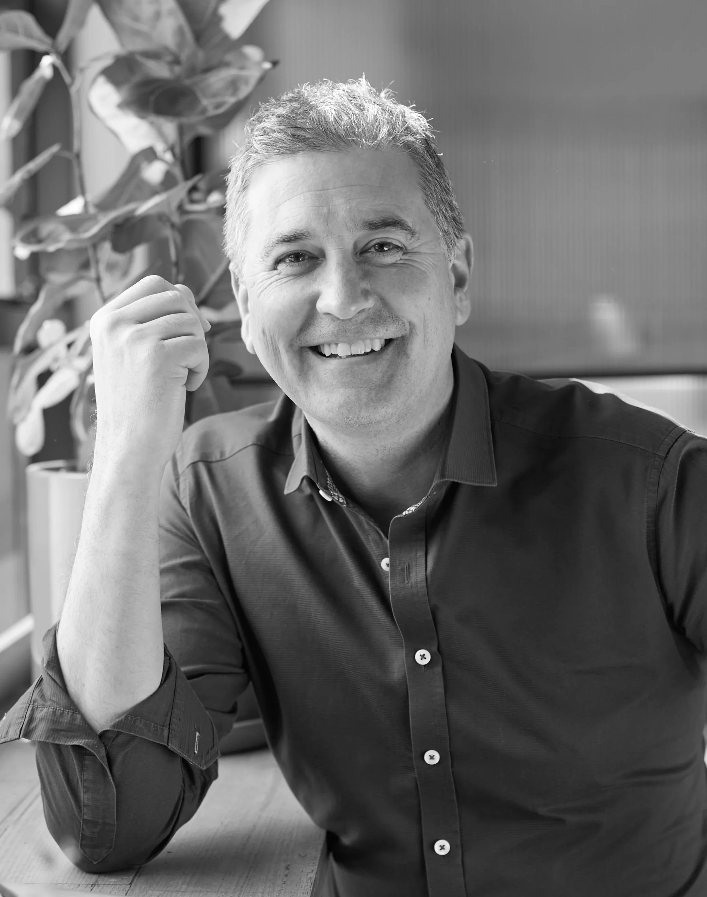 A black-and-white portrait of a middle-aged man with gray hair, sitting at a desk, wearing a light-colored shirt, with a colorful abstract artwork in the background.