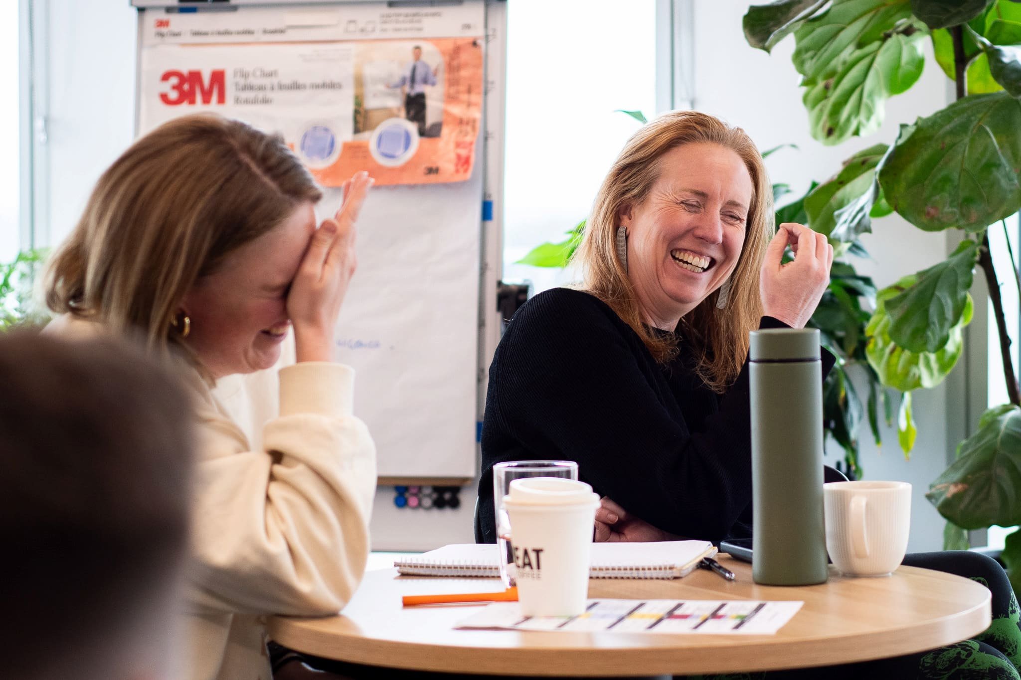 Two women sitting at a table, both laughing and smiling. One sitting on the right has red hair and is wearing a black cardigan, while the other on the left has light brown hair and is wearing a beige sweater. There are notebooks, a pen, a water bottl