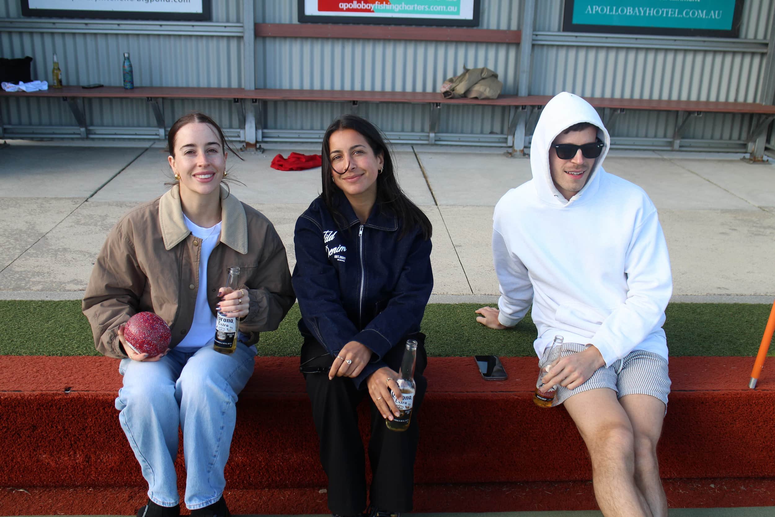 Three young adults sitting on a red bench outdoors, smiling while holding bottled drinks, with a corrugated metal wall and advertisements in the background.