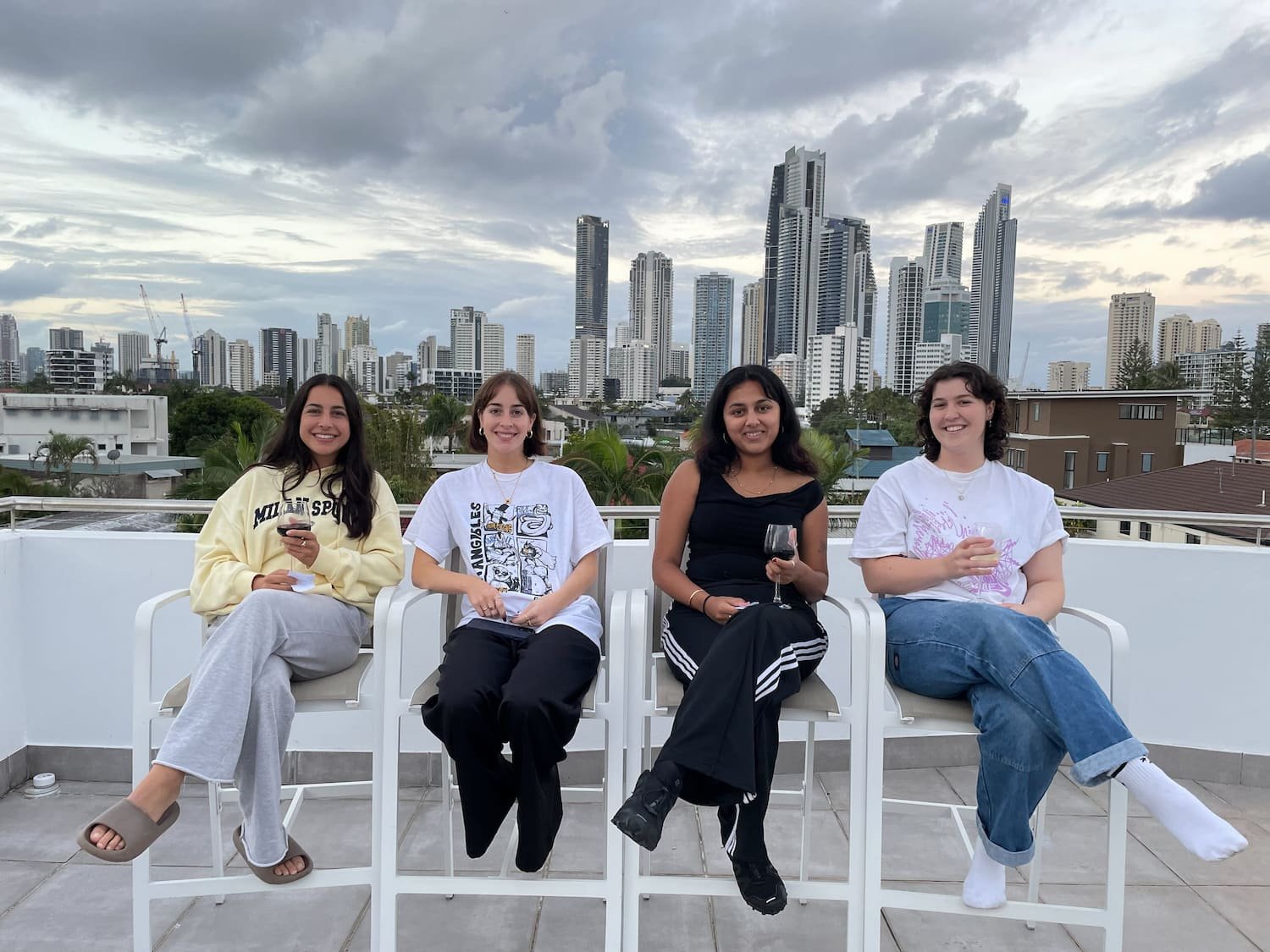 Four women sitting on white chairs on a rooftop with a city skyline in the background. They are smiling, and two of them are holding glasses of wine while one has a phone. The sky is cloudy.