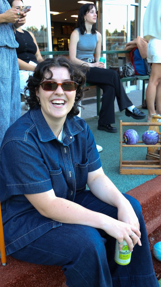 A woman with curly dark hair wearing sunglasses and a denim jacket, smiling while sitting outside. Multiple people are in the background, including a woman with dark hair in a gray sleeveless top and black pants, sitting on a bench, and other individ