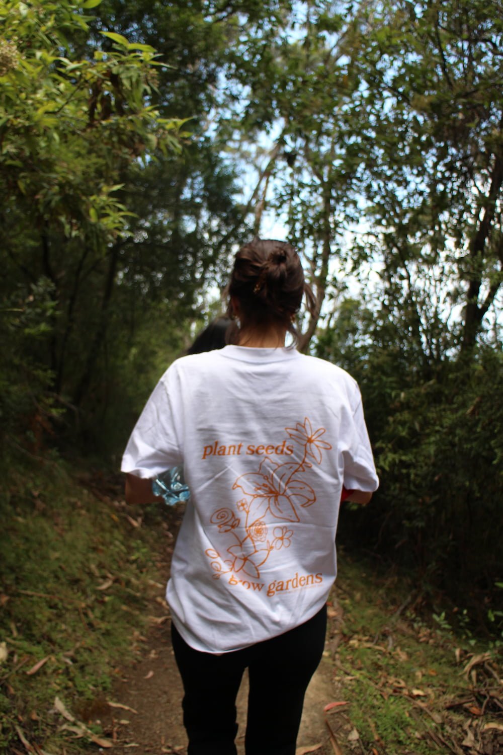 A person walking on a wooded trail wearing a white t-shirt with orange text and illustrations of flowers and the words 'plant seeds', 'grow gardens' on the back.