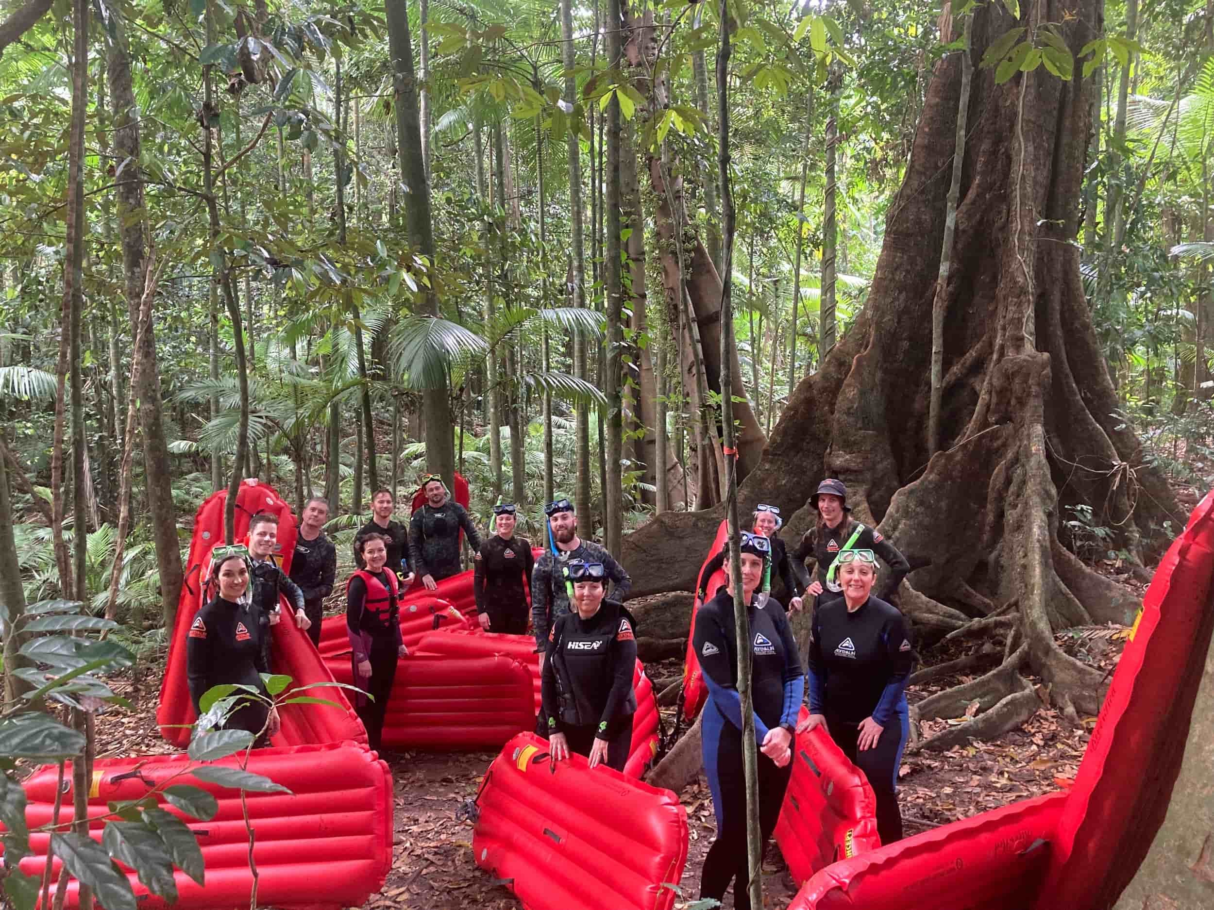 Group of people in a rainforest standing on and around red inflatable rafts, with dense green trees and a large tree with extensive roots in the background.