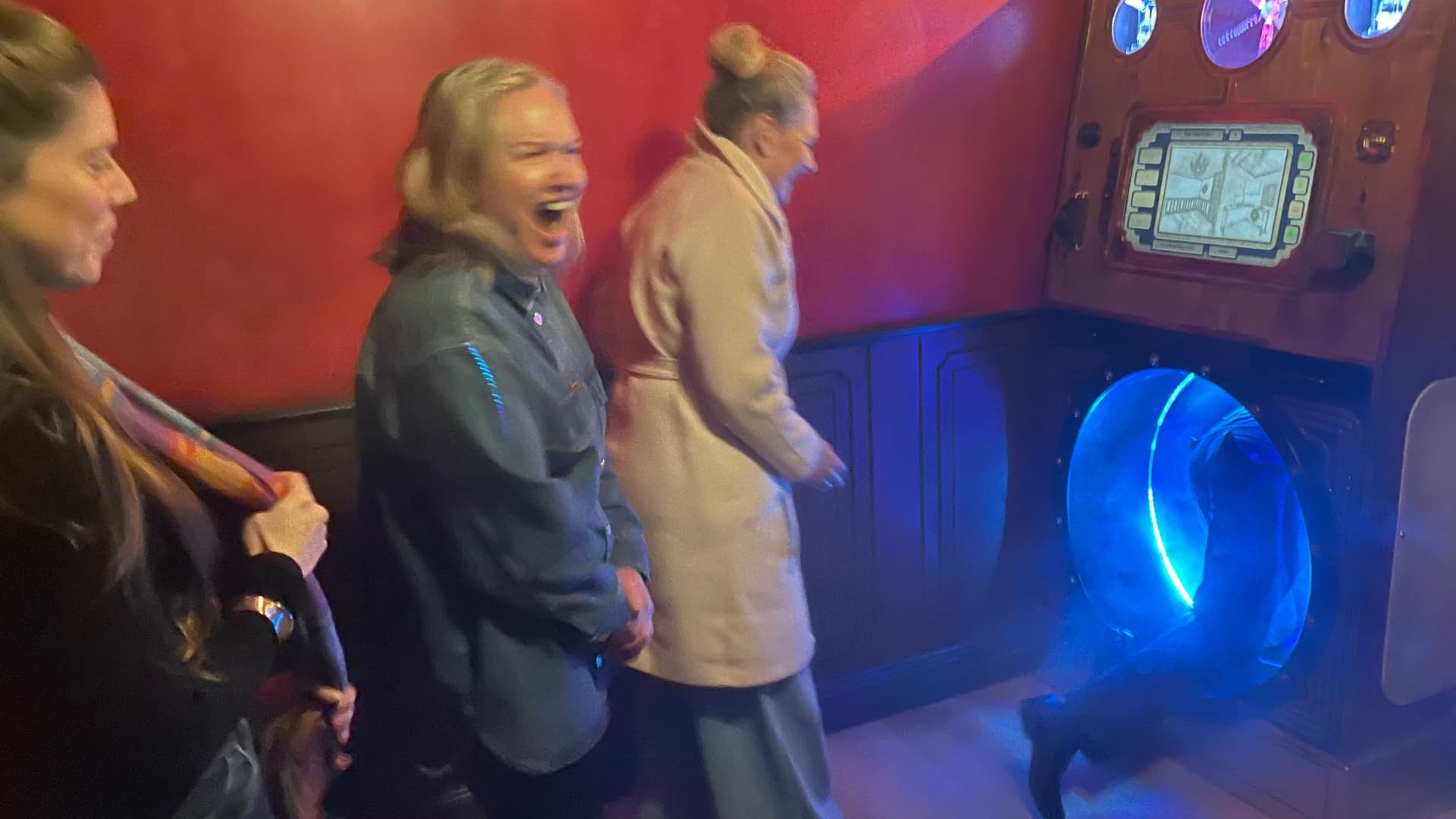 Three women standing in front of a virtual reality gaming machine, with one woman in the middle laughing or smiling, all looking toward the blue-lit VR equipment.
