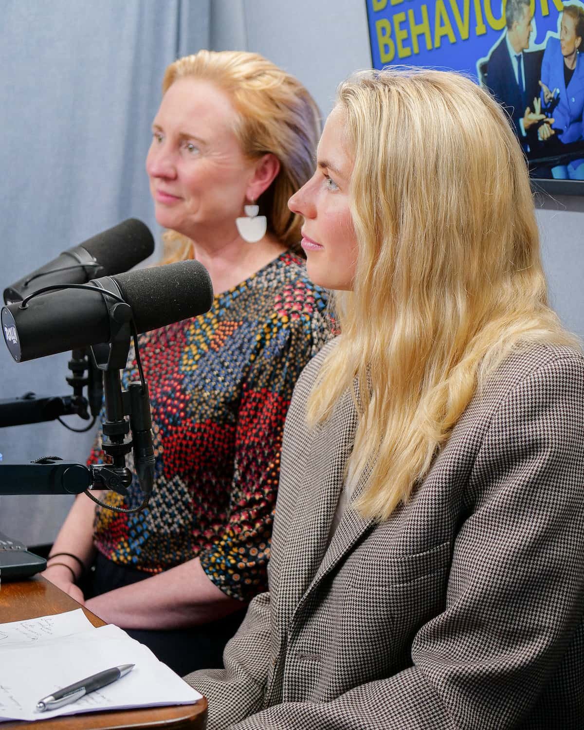 Two women sitting side by side at a table with microphones, appearing to participate in a discussion or podcast, with a TV screen in the background showing two people and the text "Behavior"