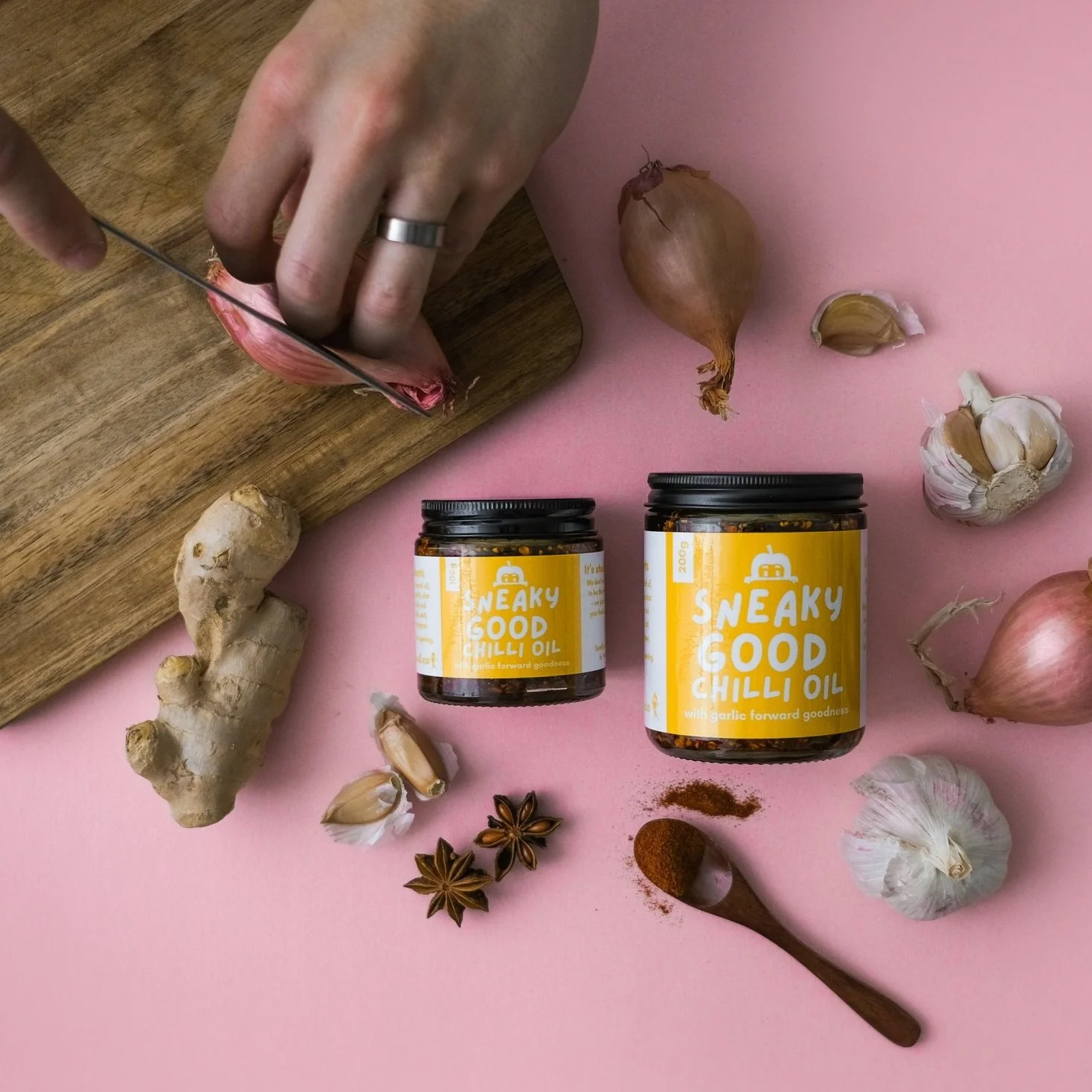 Hand slicing a shallot on a wooden cutting board surrounded by jars of Sneaky Good Chilli Oil, garlic bulbs, ginger, star anise, and a wooden spoon with chili powder on a pink background.