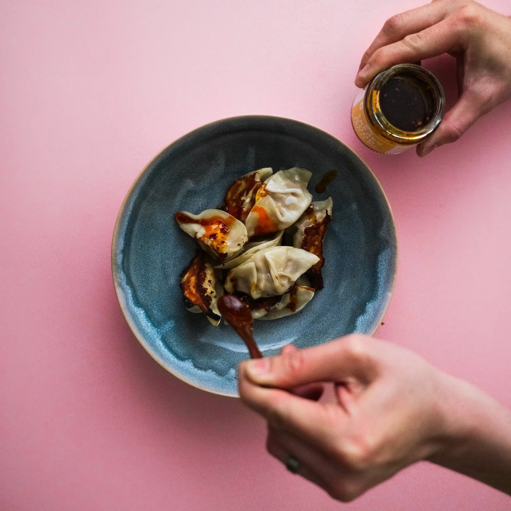 Hands holding a jar and spoon, adding sauce to dumplings in a blue bowl, placed on a pink surface.