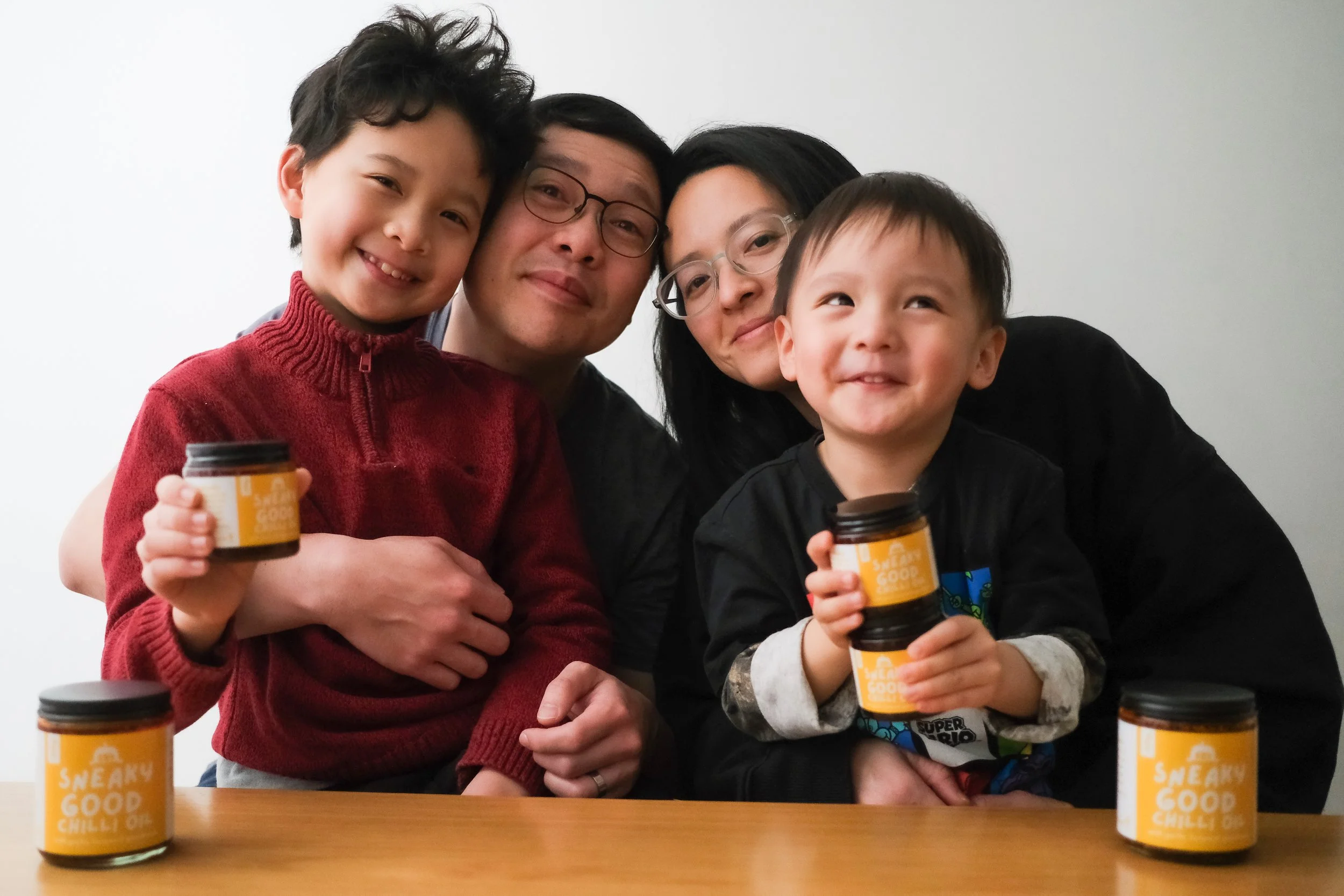 Family of four smiling, holding jars labeled 'Sneaky Good Chili Oil,' some jars are on the table.