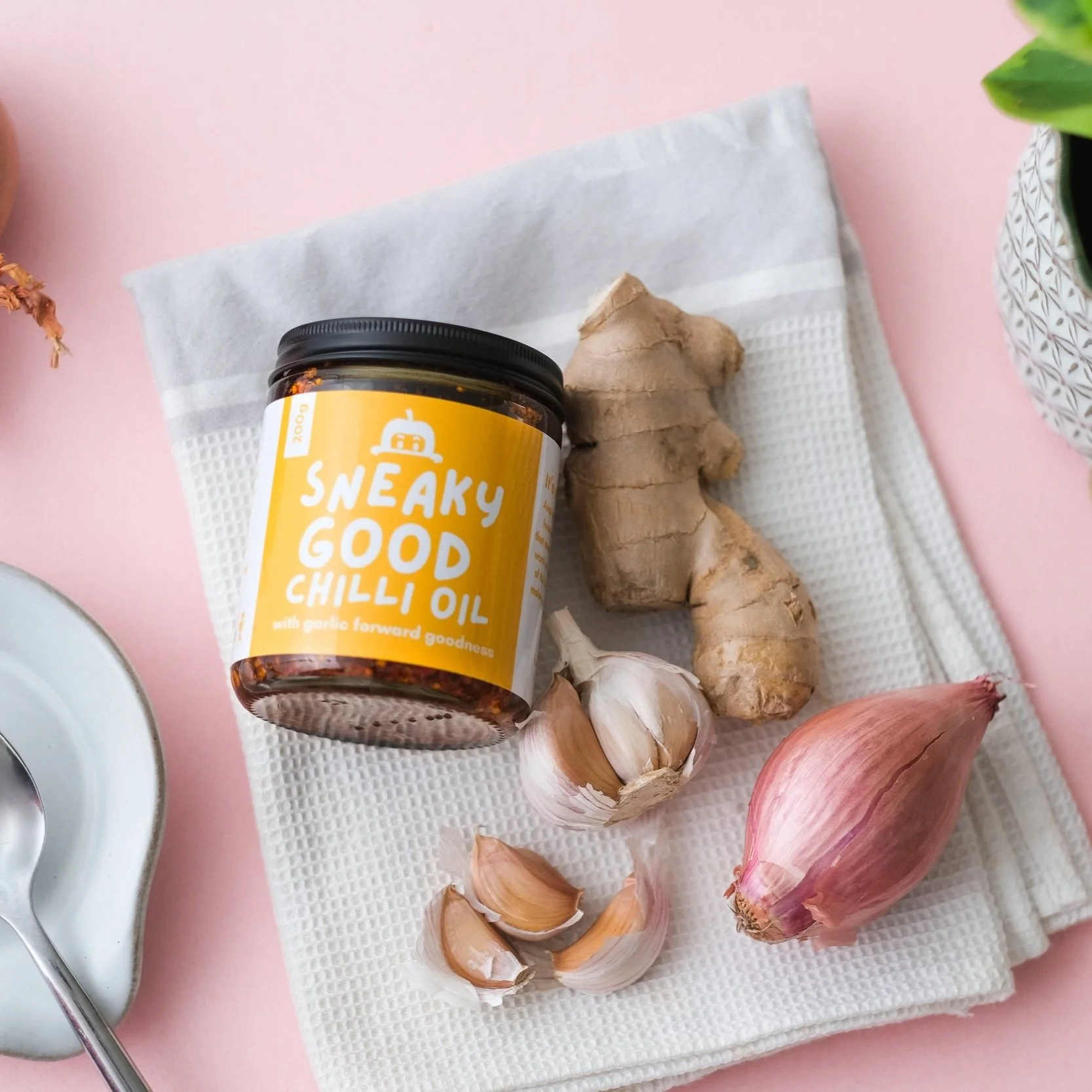 Jar of Sneaky Good Chilli Oil with garlic, ginger, and shallot on a cloth, placed on a pink background.