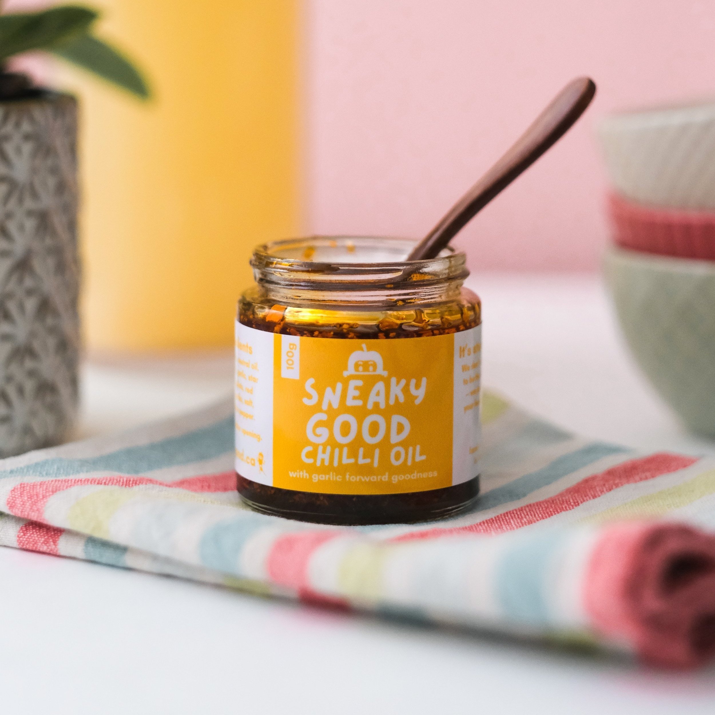 Jar of Sneaky Good Chilli Oil with spoon on colorful cloth, decorative plant, and bowls in background.