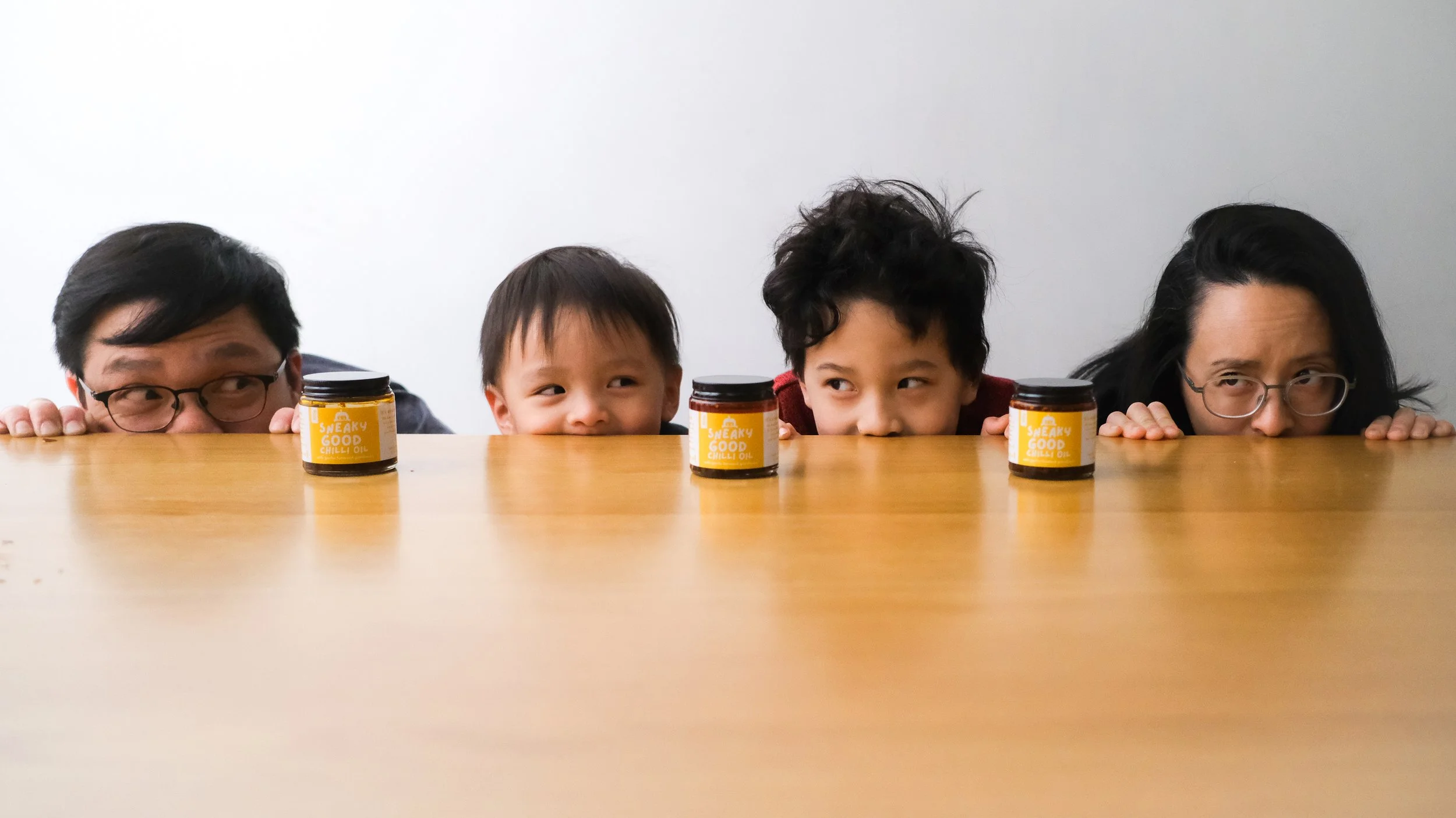 Four people peeking over a table with jars labeled 'Sneaky Good Chilli Oil' on it.