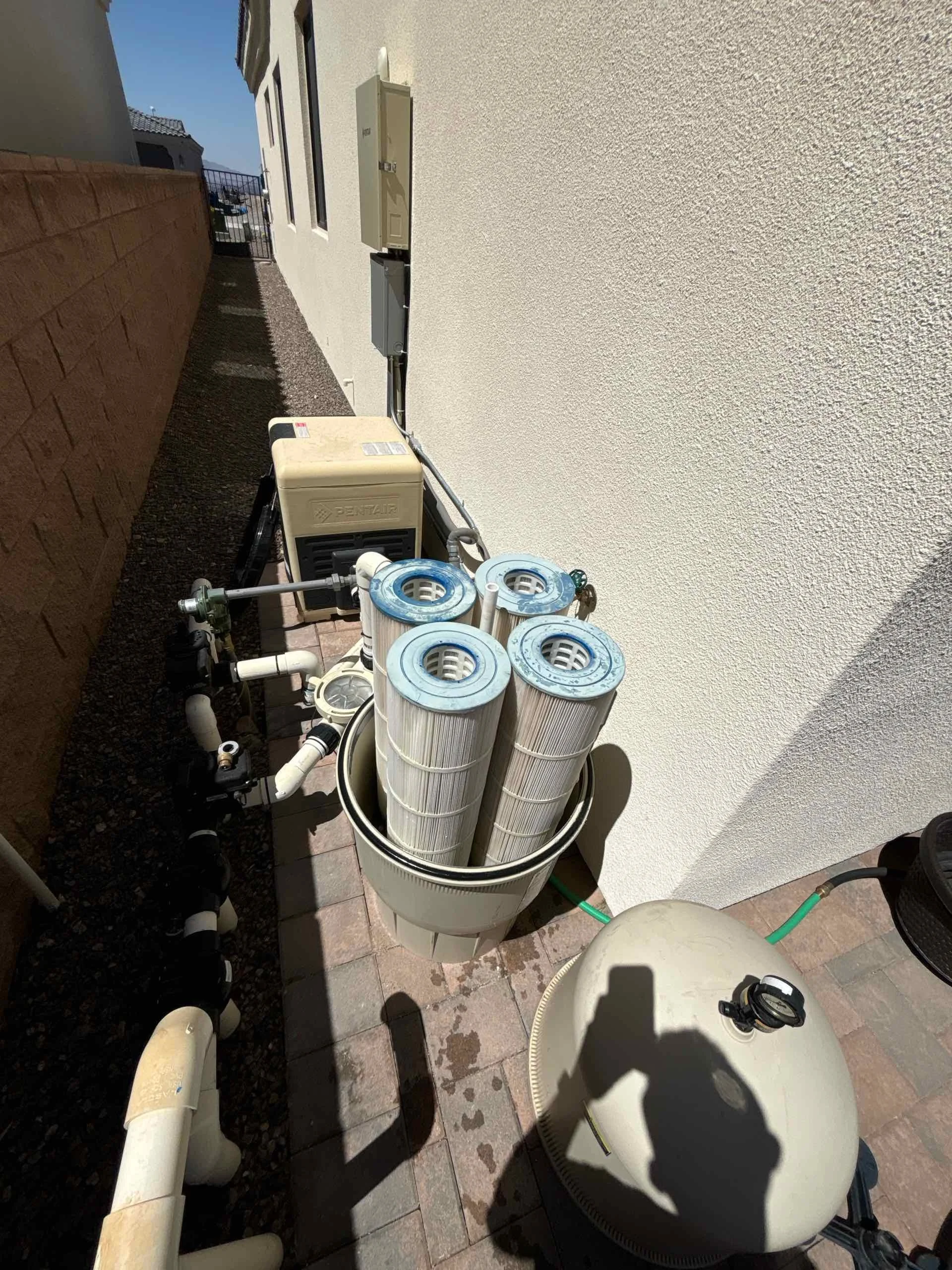 Pool filter cartridges, pump, and filtration system on a patio next to a white building wall.