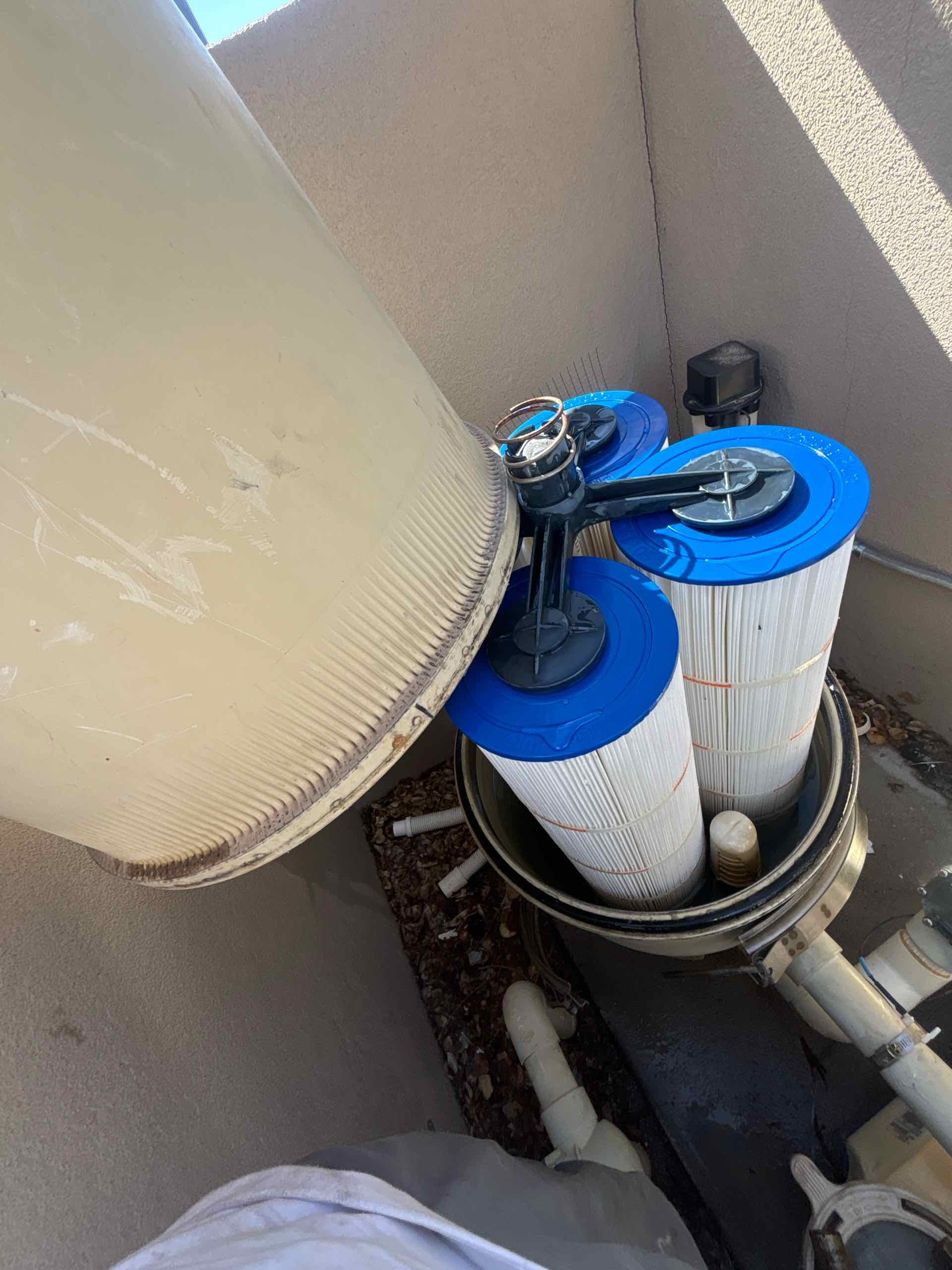 Pool filter system with two large cylindrical filters and a pump, installed on a concrete patio with a beige wall in the background.