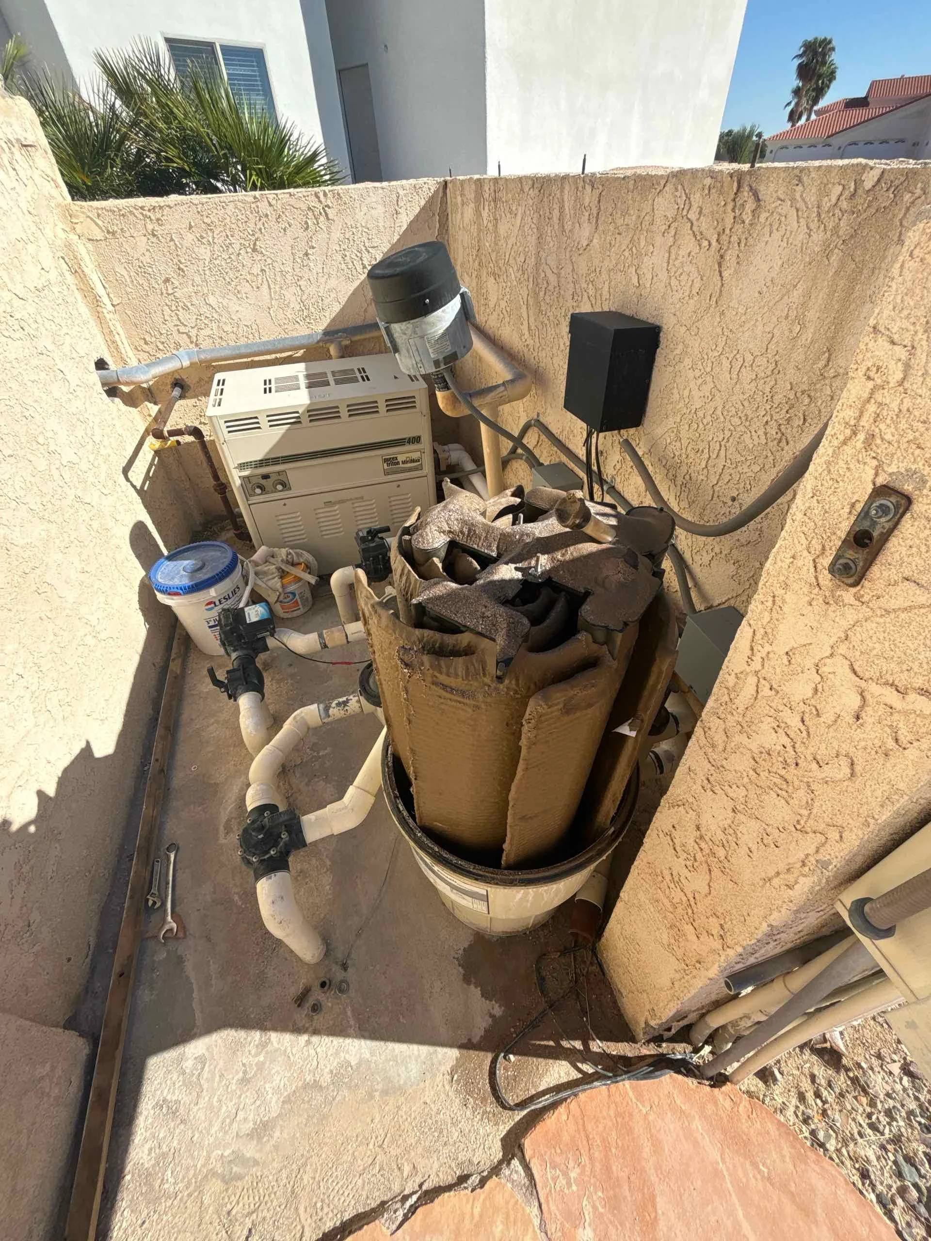 A small outdoor utility space with a pool filter, pump, and heater surrounded by beige stucco walls, with piping, a bucket, and some tools visible.