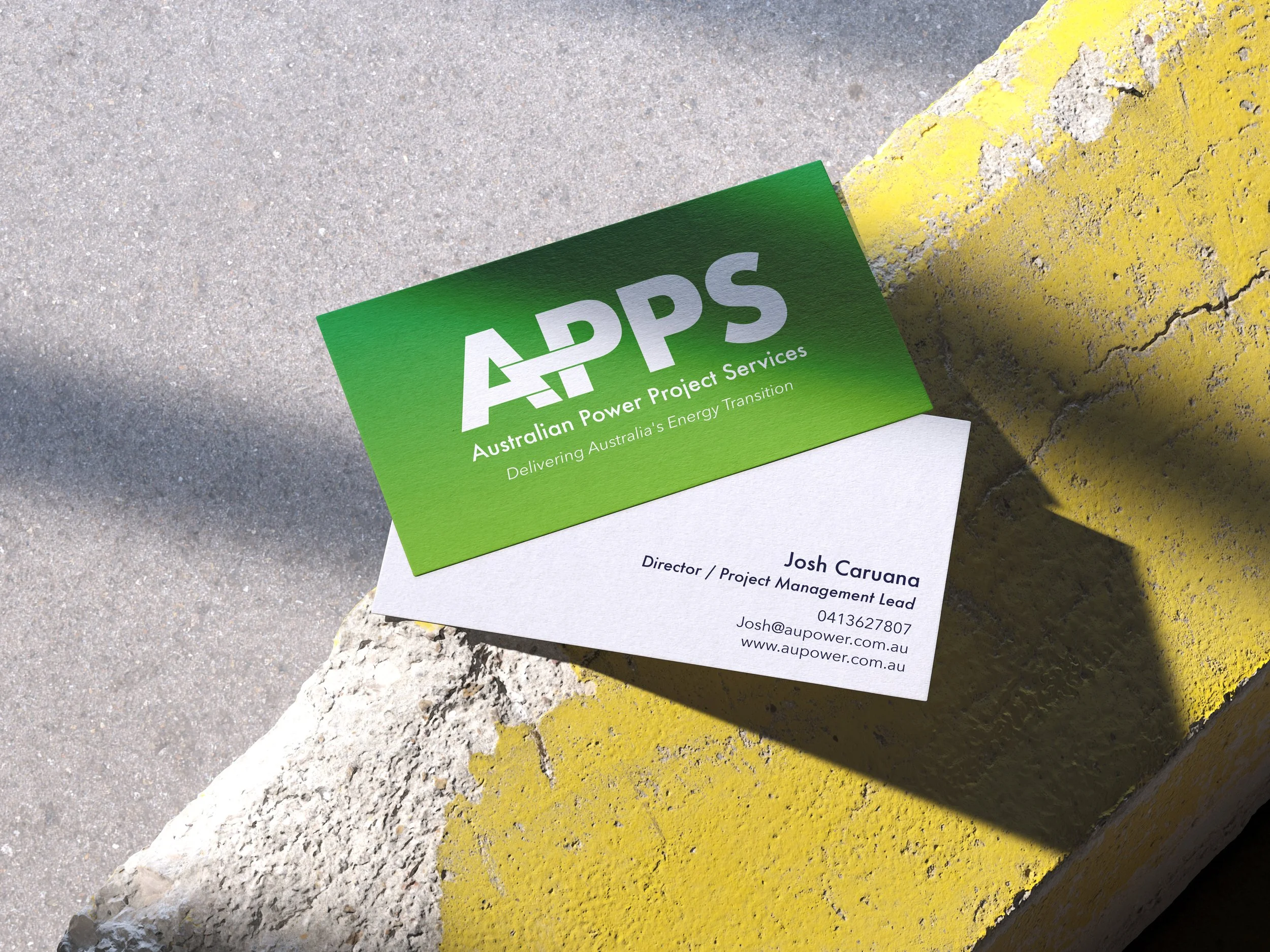 A business card for Australian Power Project Services (APPS), placed on a yellow and white curb with a concrete sidewalk nearby. The card features green and white colors, with the company logo, contact information for Josh Caruana, and the website addresses.
