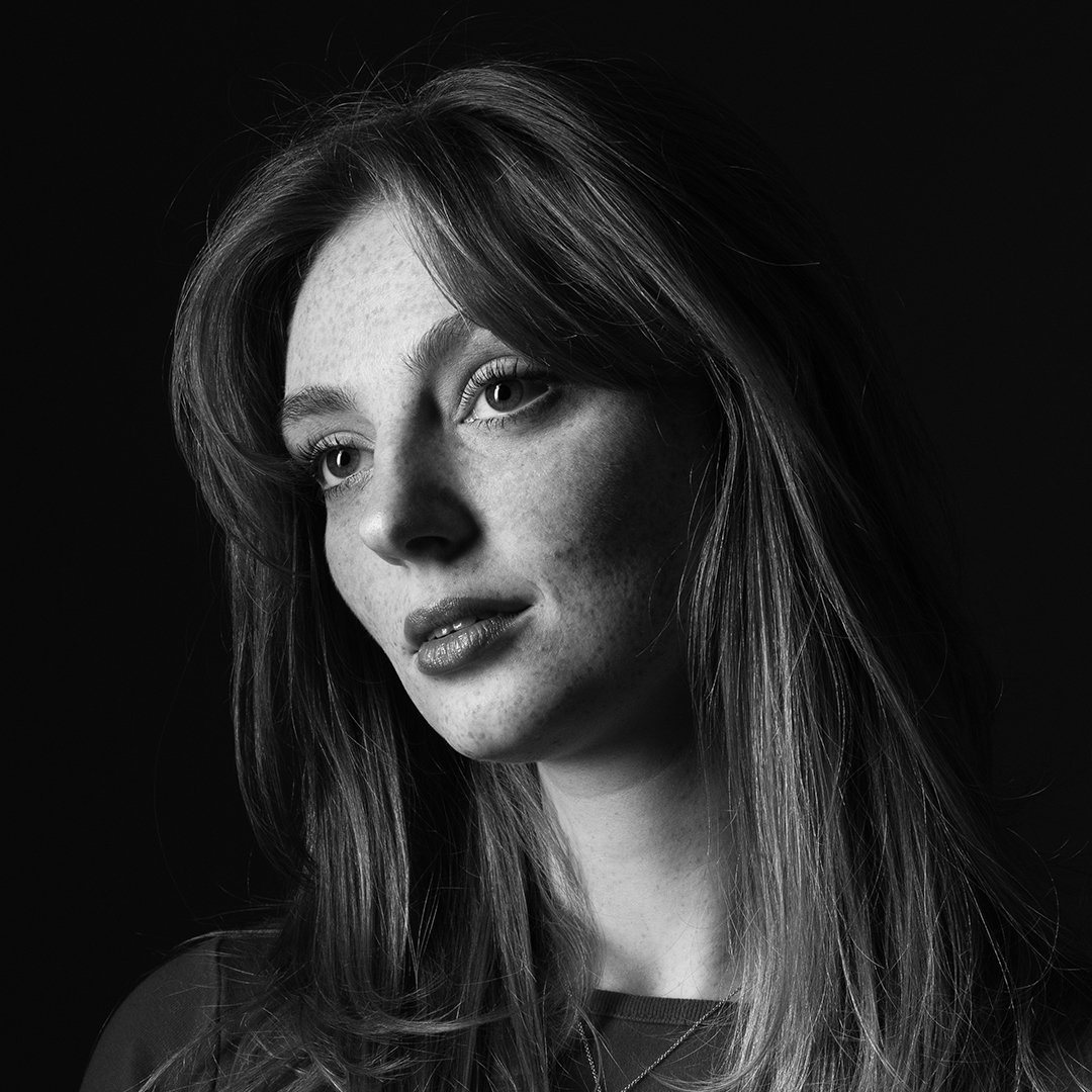 Black and white portrait of a woman with long hair, looking slightly away from the camera, with natural makeup and freckles on her face, against a dark background.