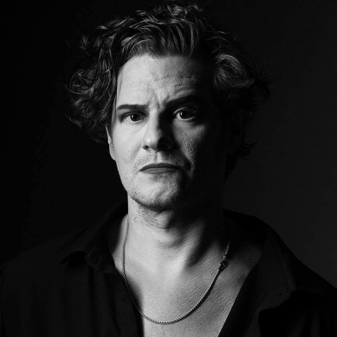 Black and white portrait of a man with wavy hair, wearing a dark shirt and a chain necklace, against a dark background.