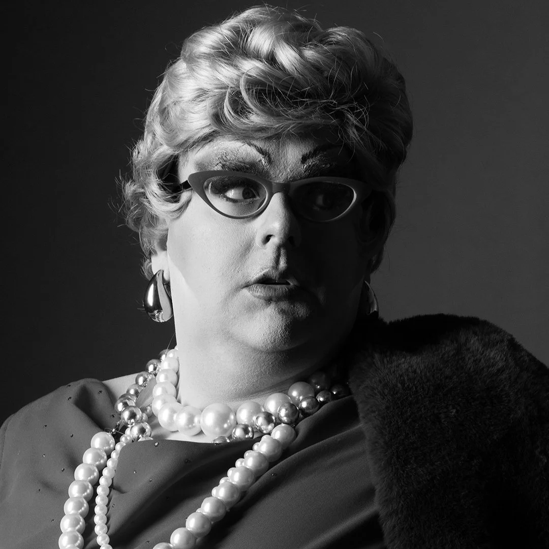 Black and white portrait of a person with short, curly hair wearing glasses, earrings, and multiple pearl necklaces, dressed in elegant clothing with a fur collar.