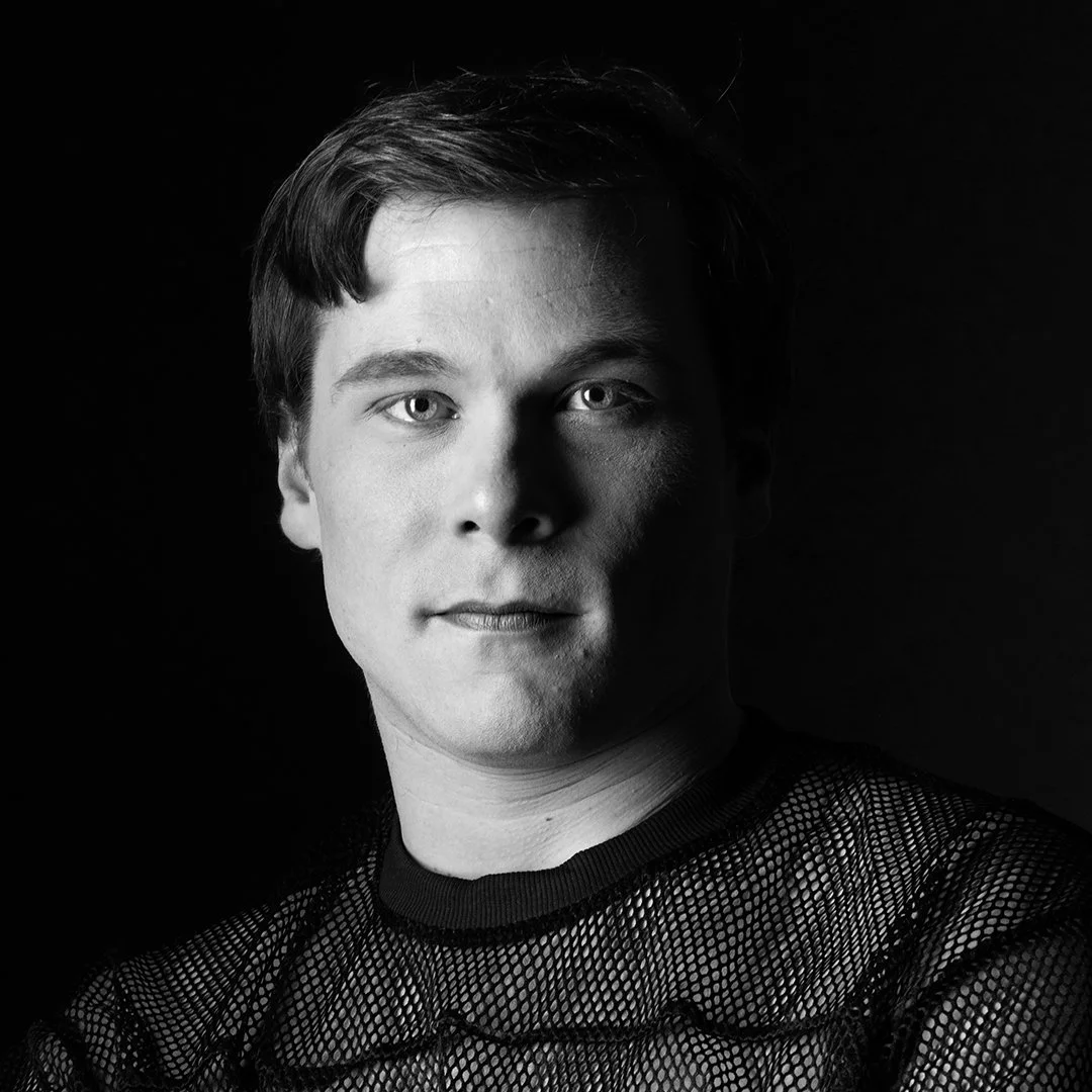 Black and white portrait of a young man with short hair, wearing a mesh top, against a dark background.