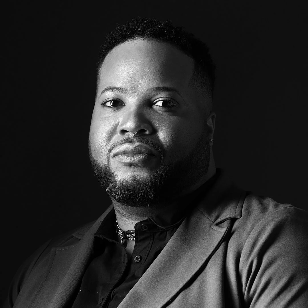 Black and white portrait of a man with a beard, wearing a suit and a necklace, against a dark background.