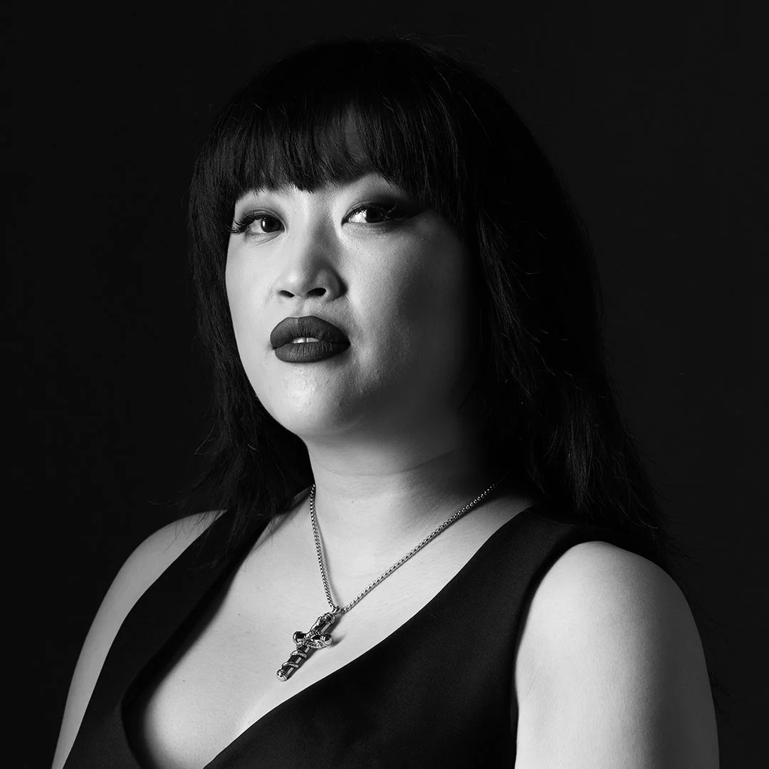 Black and white portrait of a woman with dark hair and bangs, wearing dark lipstick, a black top, and a necklace with a cross pendant, against a dark background.