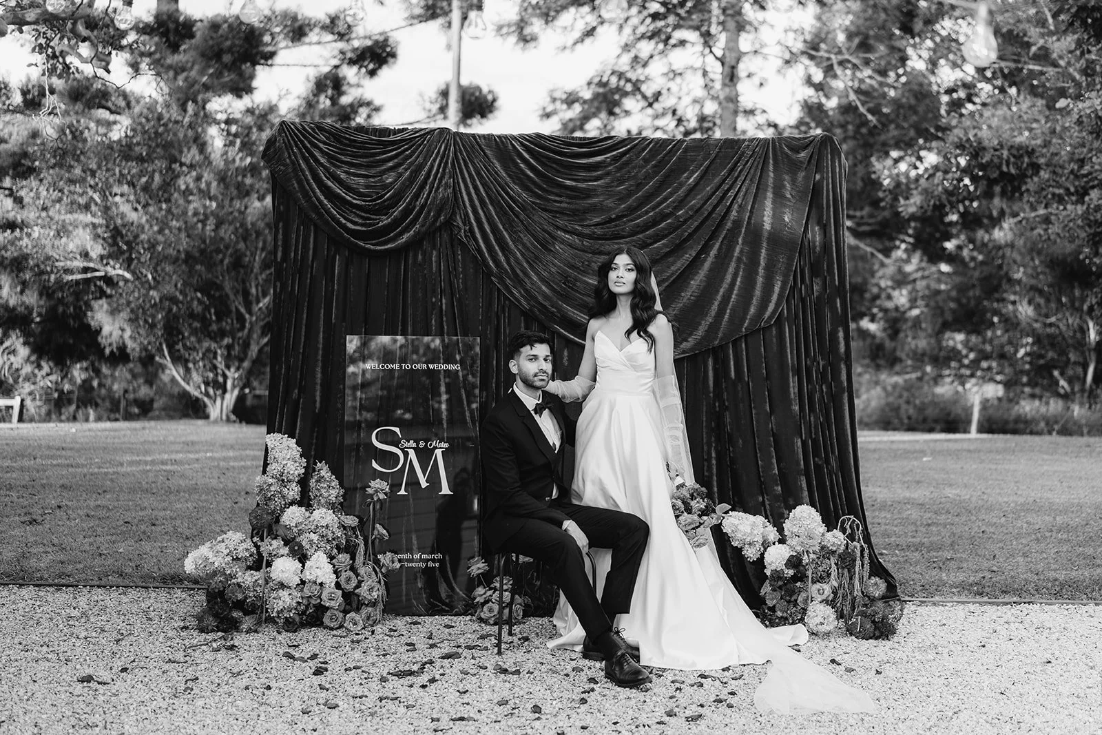 A couple dressed in wedding attire posing in front of a black draped backdrop with floral arrangements on the ground, outdoors with trees in the background.